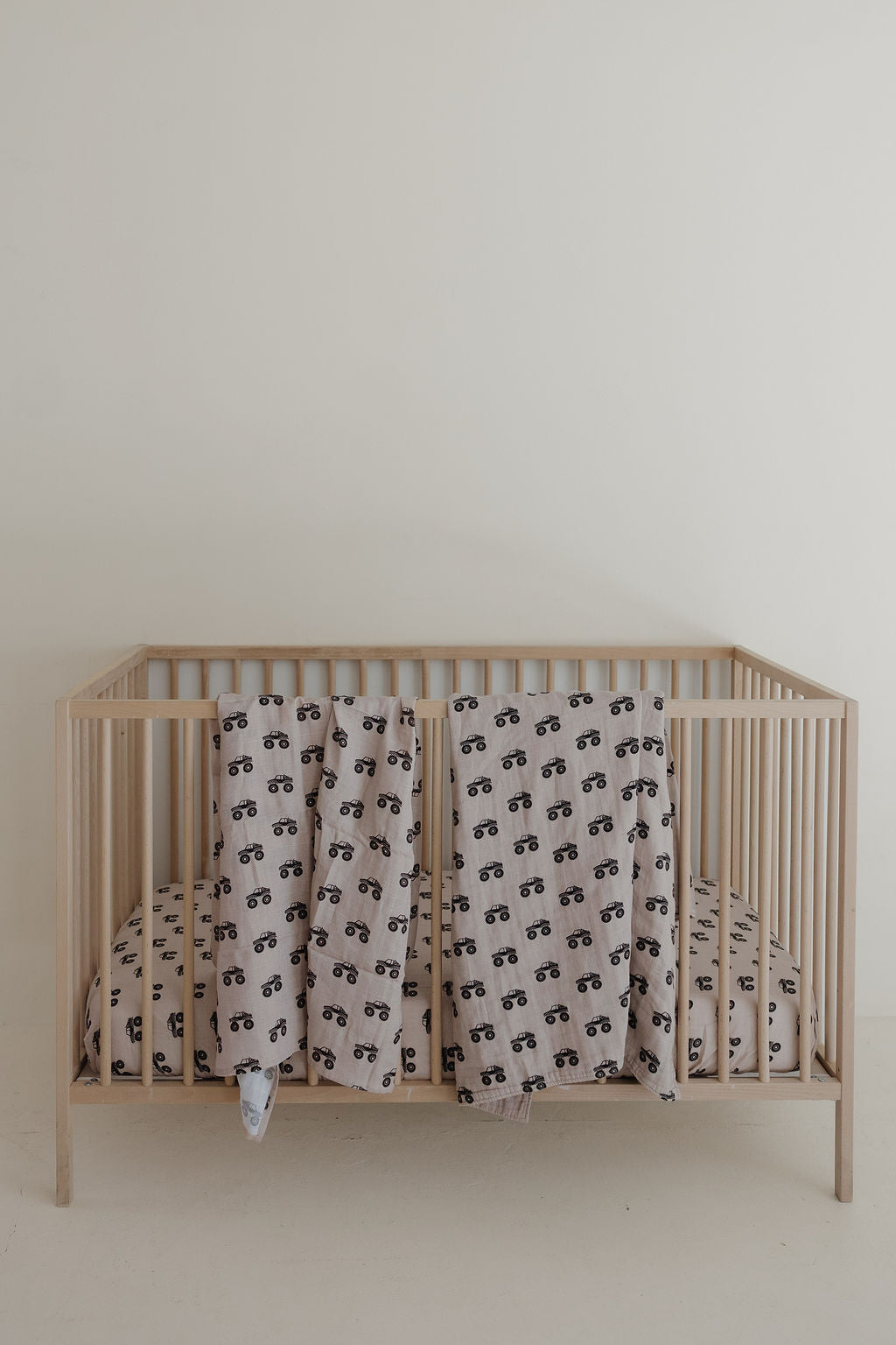 A light wood baby crib against a plain white wall, with matching bedding and two forever french baby Muslin Swaddle | Monster Truck in ultra-soft cotton, draped over the side, all featuring a black monster truck print on a light beige background.