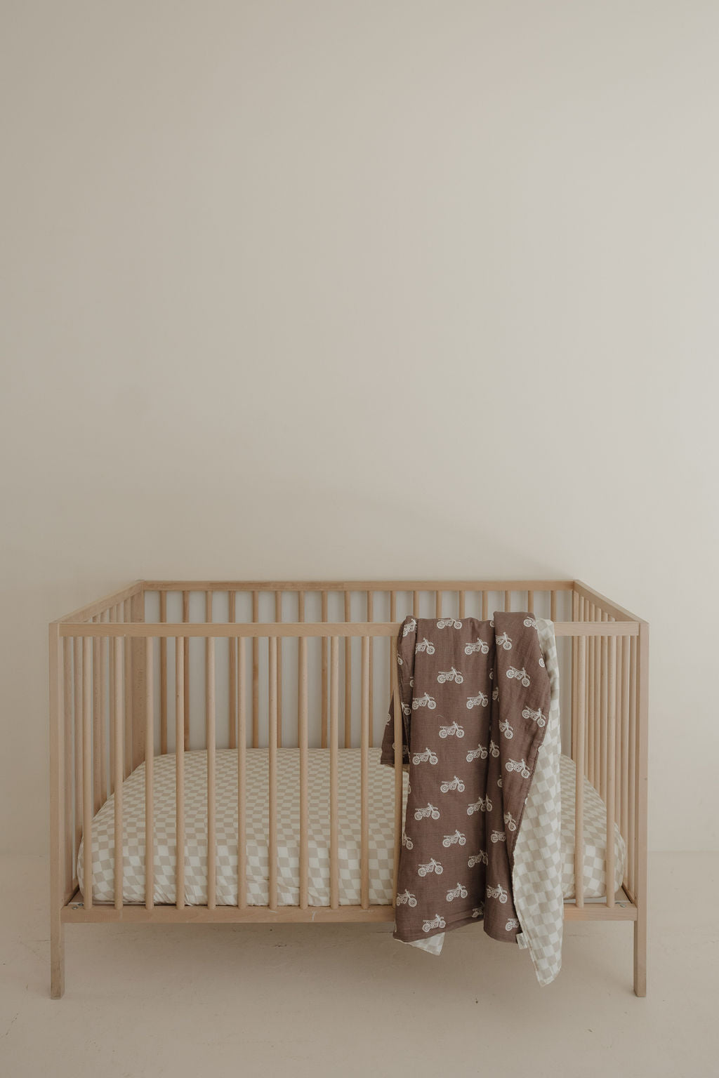 A minimalist wooden crib with a patterned mattress and a baby nursery quilt, plus the forever french baby Muslin Quilt | Dirt Bike & Checkerboard draped over the side, set against a plain beige wall.