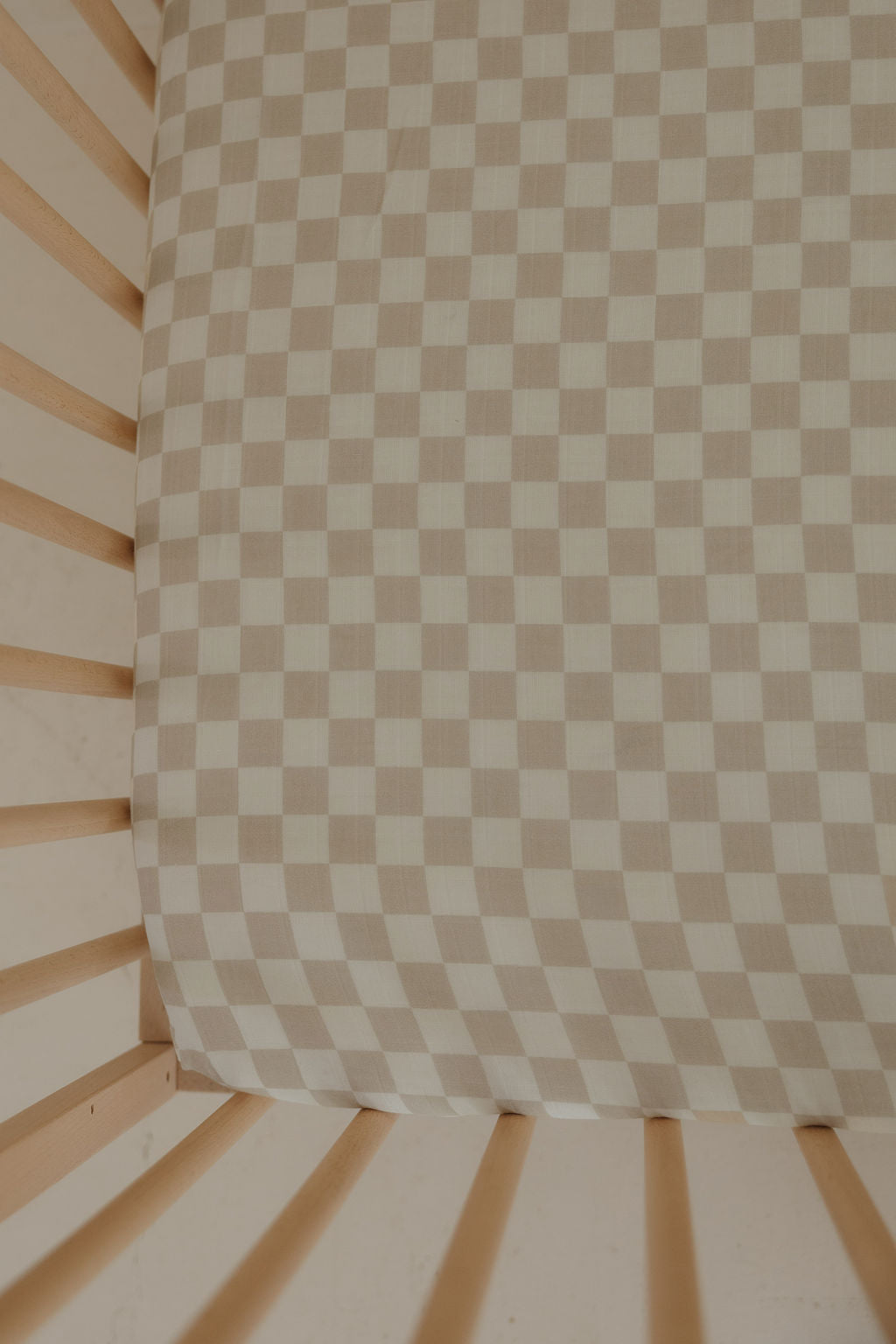 The Muslin Crib Sheet | Checkerboard by forever french baby features a soft beige and white checkerboard design, pairing perfectly with light wooden crib slats for cozy, stylish baby bedding.