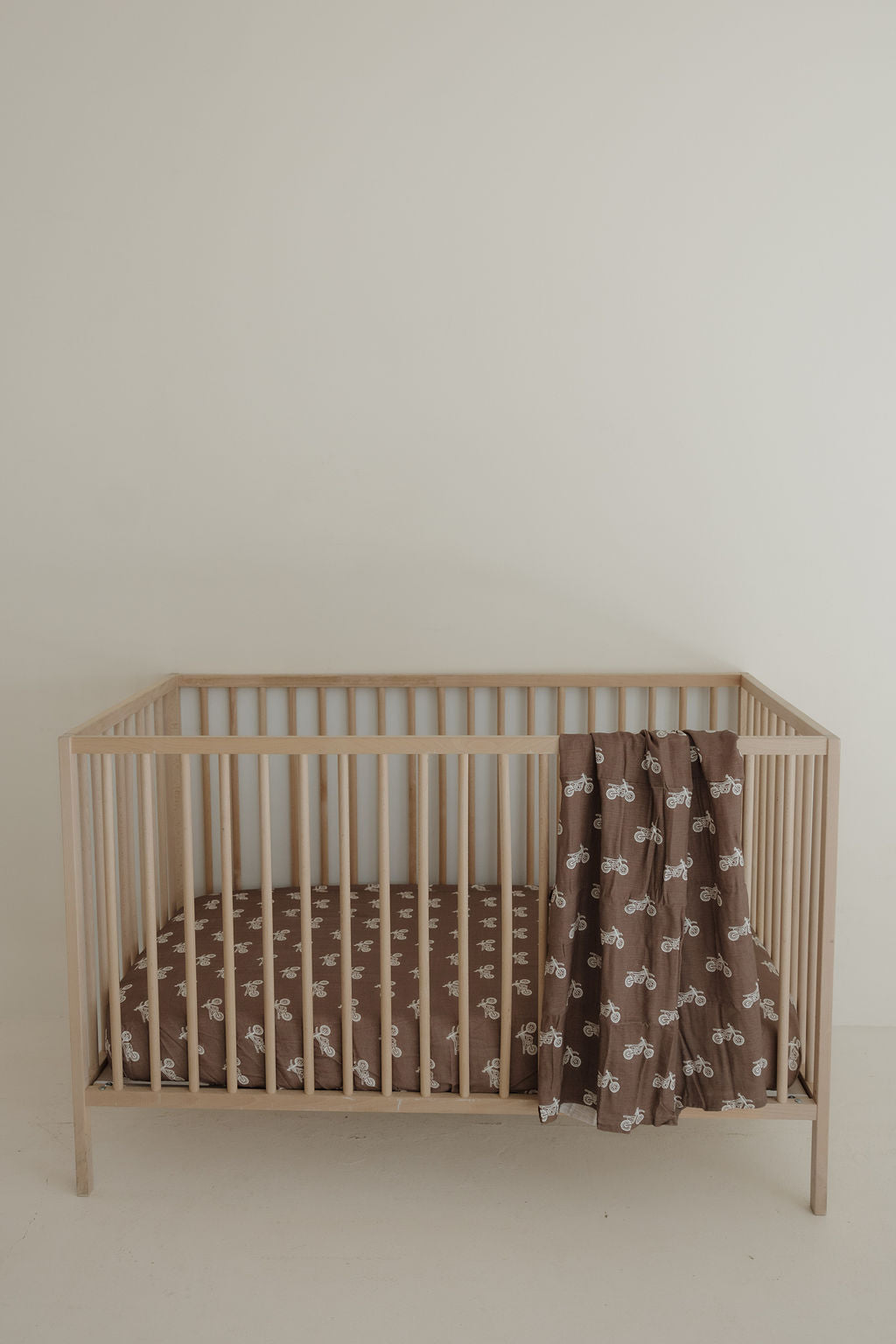 A light wooden baby crib features the forever french baby Muslin Crib Sheet in the Dirt Bike design, with matching brown nursery bedding and a brown blanket with a white giraffe pattern, all set against a plain off-white wall.