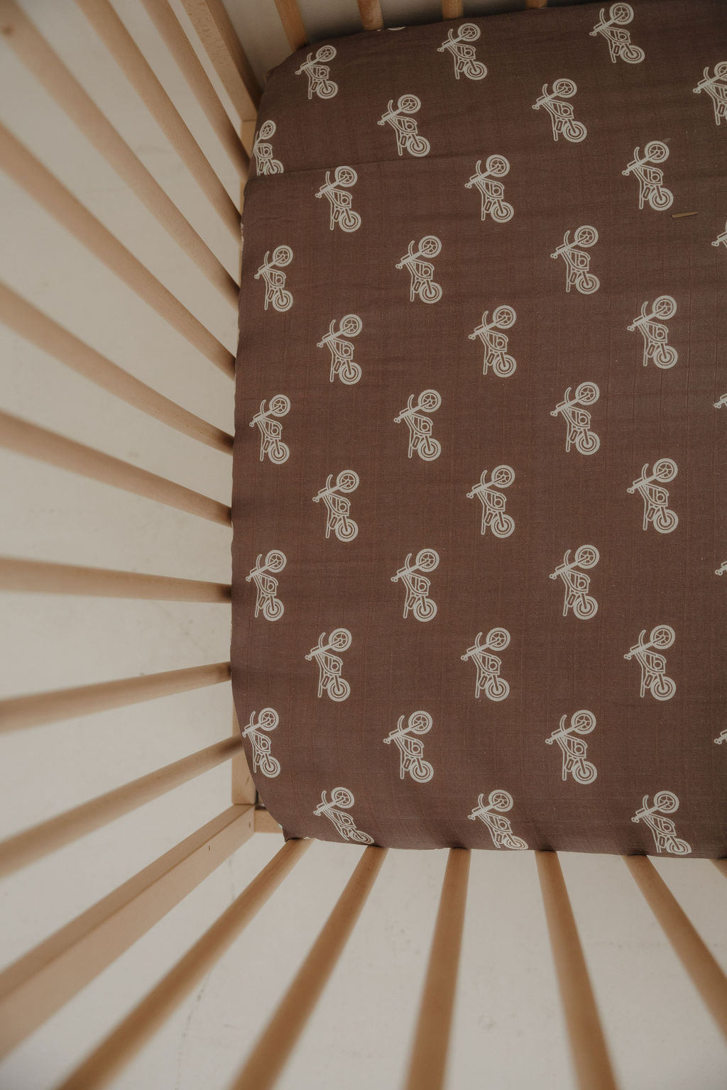 A crib with light wooden slats and nursery bedding, including the forever french baby Muslin Crib Sheet | Dirt Bike in brown with a white bear riding a bicycle line drawing pattern.