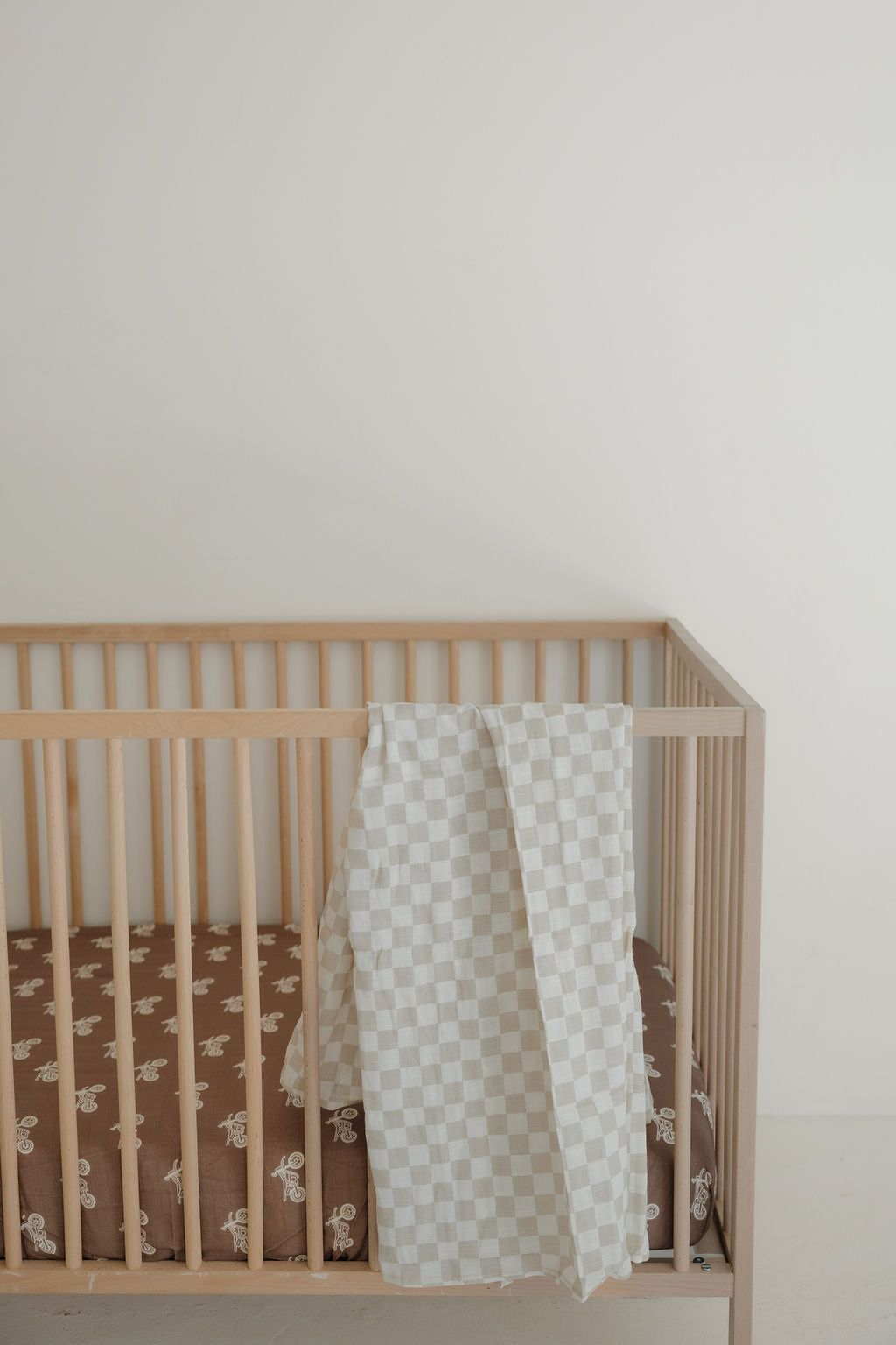 A light wooden crib with a brown mattress featuring white patterns and a forever french baby Muslin Swaddle | Checkerboard draped over the side, set against a plain white wall.