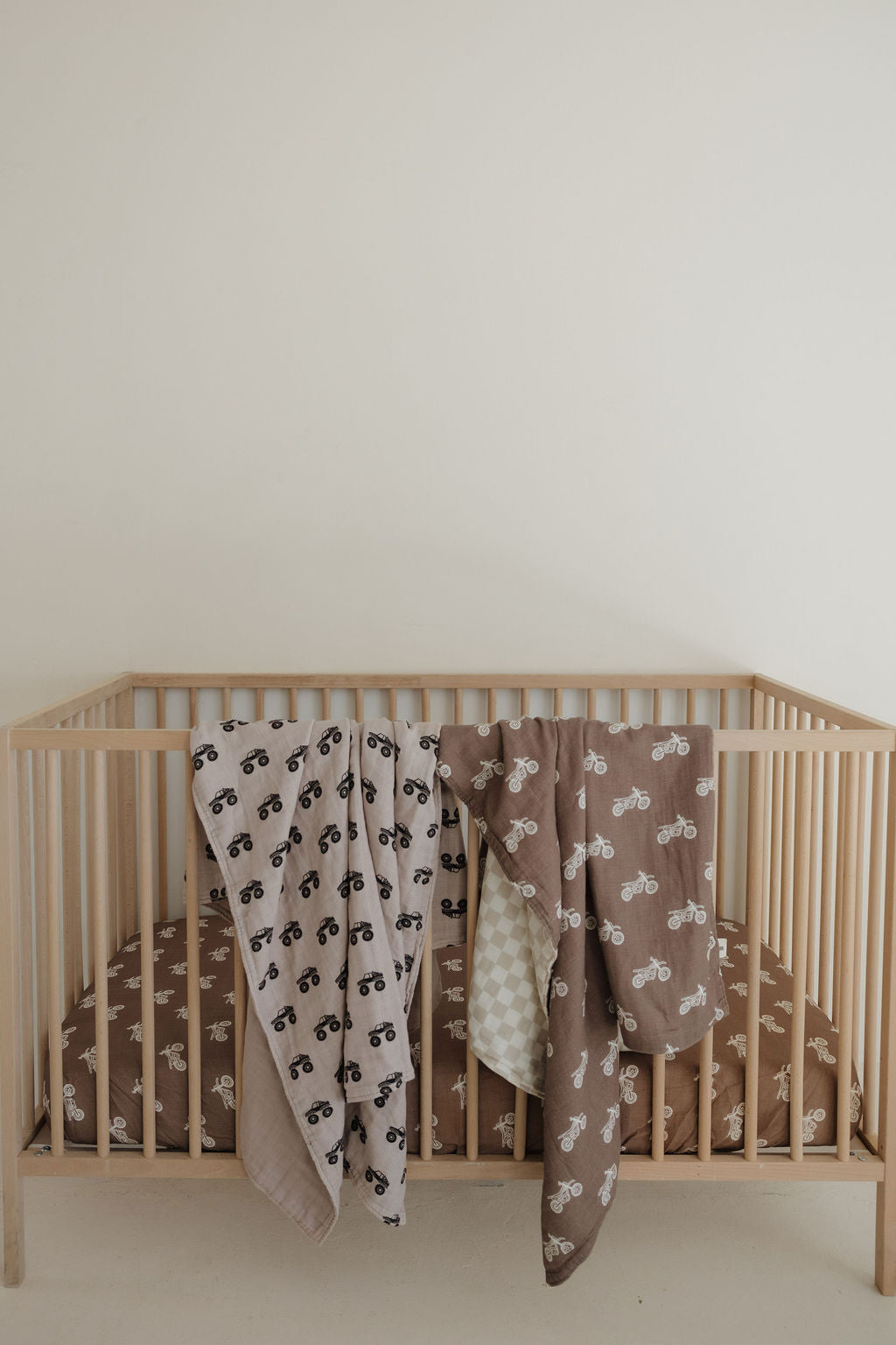 A light wooden crib with a brown fitted sheet and two muslin blankets, including the forever french baby Muslin Quilt | Dirt Bike & Checkerboard, draped over the side, set against an off-white wall.