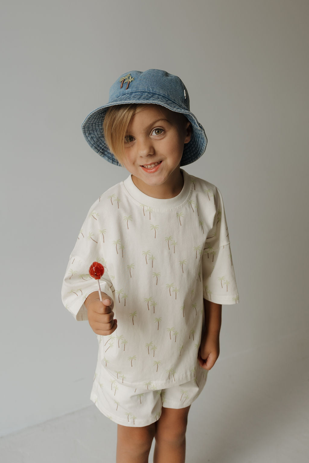 A smiling child wears the forever french baby Child Short Set | Palm—cream-colored cotton kids’ clothing adorned with small palm tree prints—and a blue bucket hat, holding a red lollipop against a plain light background.