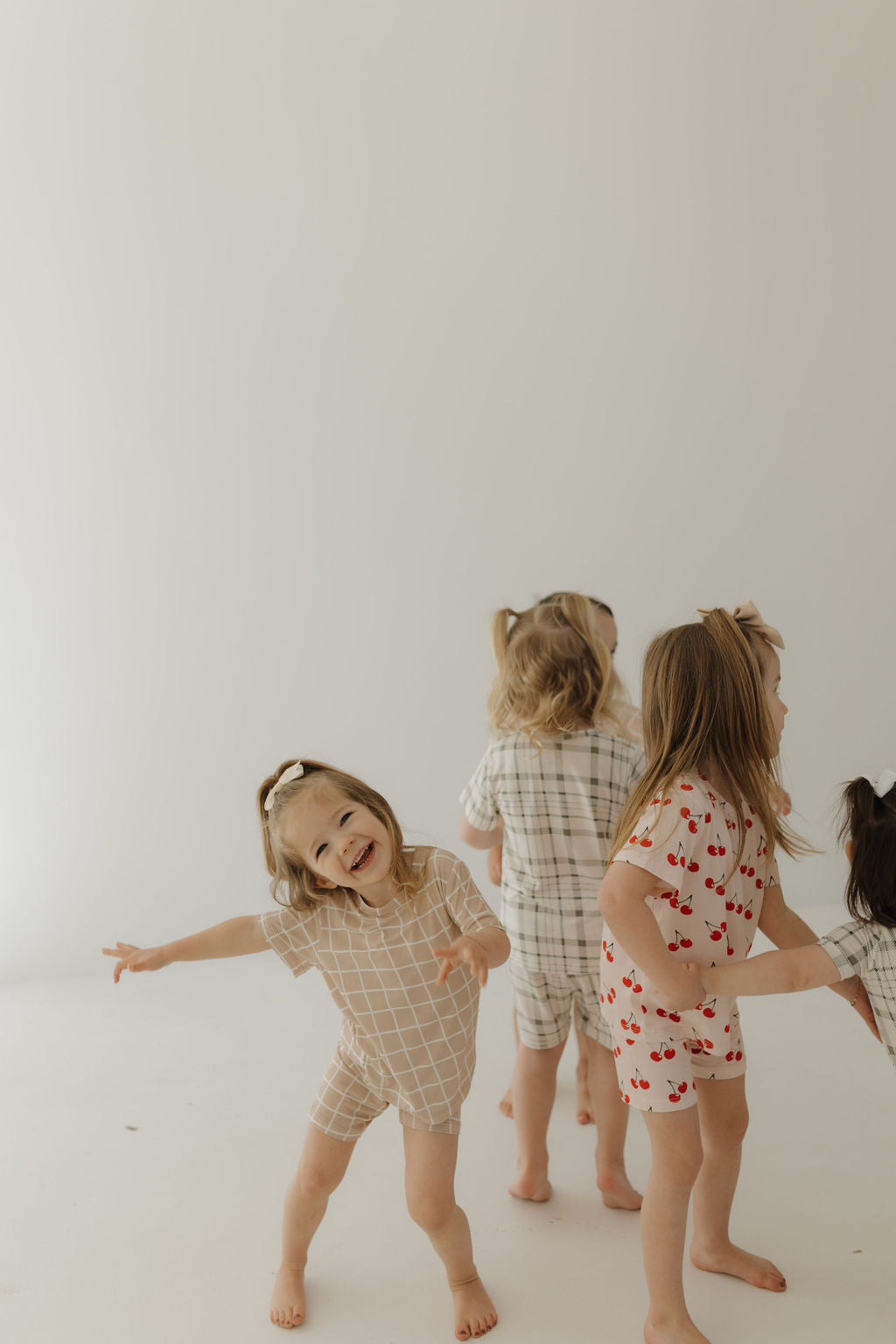 In a bright, minimalist room, children in patterned, breathable clothing play together. One child smiles and strikes a playful pose in forever french baby's Bamboo Short Set | Cherry Orchard, while others clad in hypo-allergenic outfits engage with each other, mostly facing away.