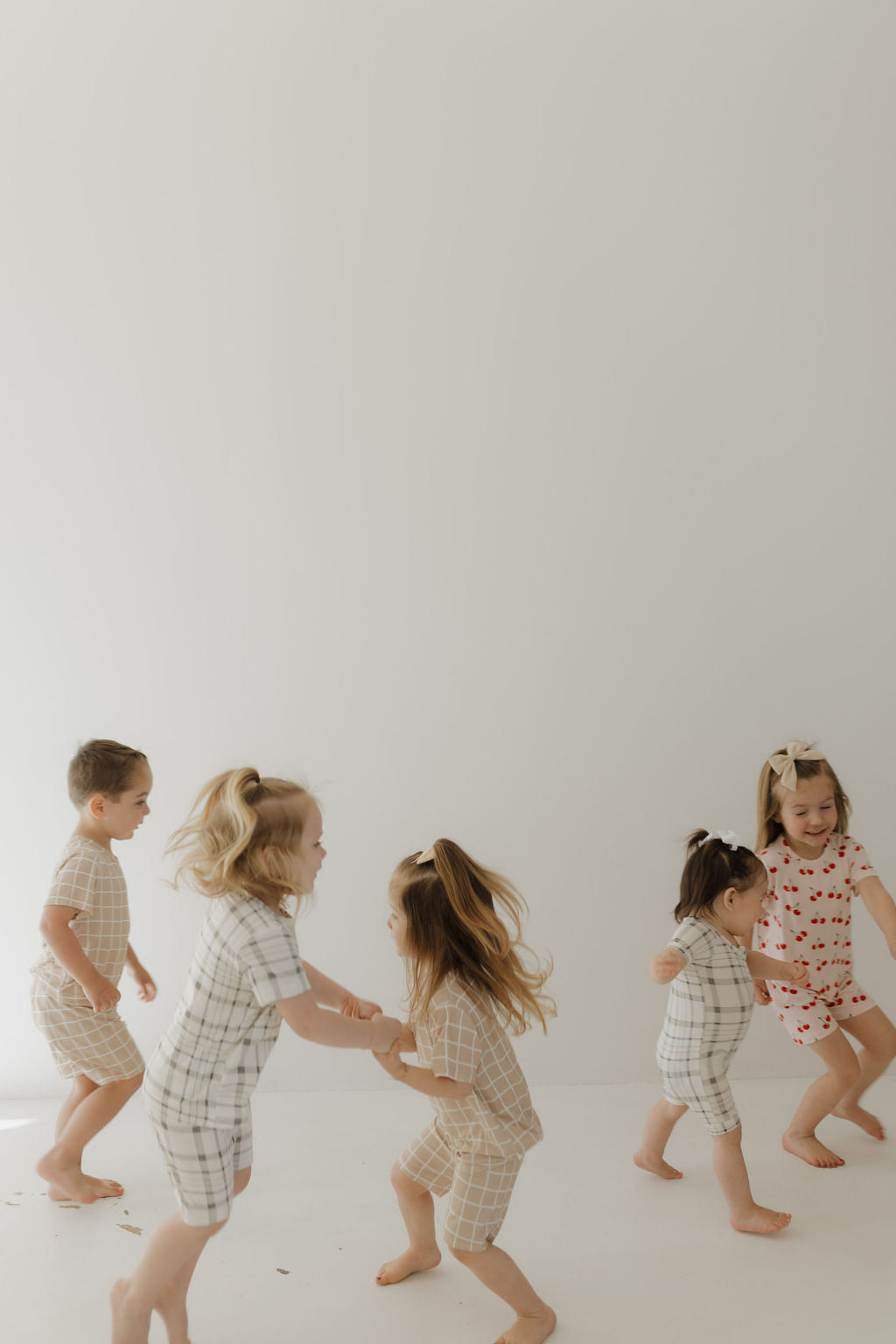 Five children joyfully play in a bright, minimalistic space wearing matching outfits. Two girls in the "Bamboo Short Set | Cherry Orchard" by forever french baby hold hands while running, as the others move energetically in their breathable fabric attire.