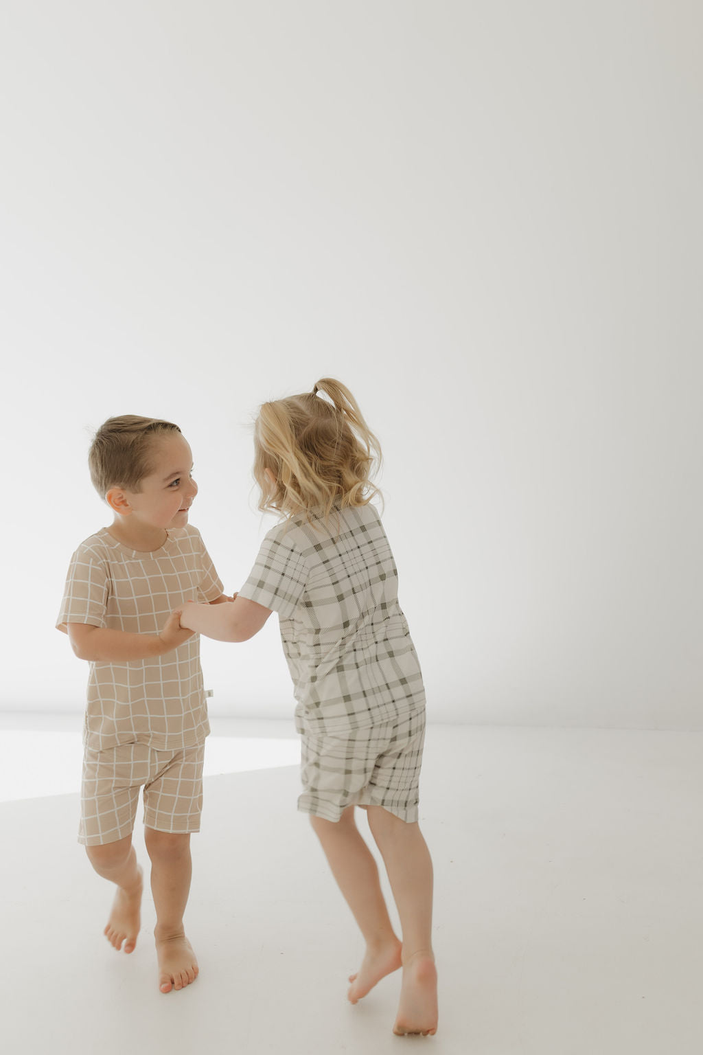 In a bright, minimalistic space, two children play together. One wears a beige Bamboo Short Set | Grid from forever french baby, while the other dons a gray plaid dress made from breathable, hypo-allergenic fabric. They hold hands and smile as they move around the light-colored background.