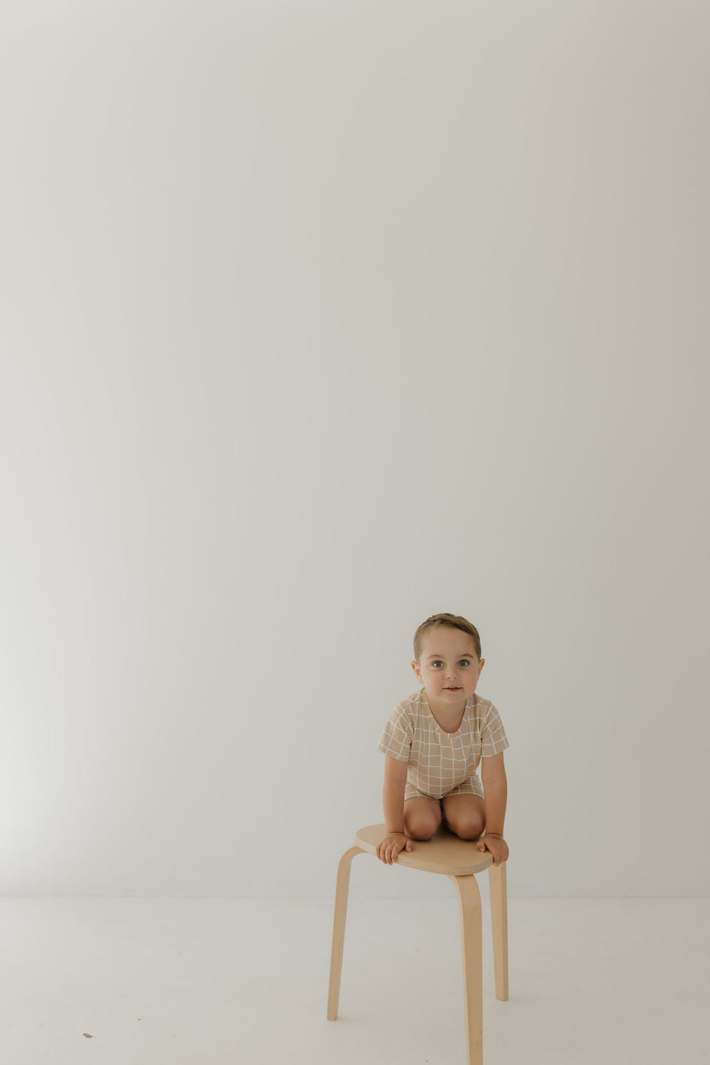A child with short hair sits cross-legged on a small wooden stool against a plain white background, wearing the Bamboo Short Set | Grid by forever french baby and appearing with a neutral expression.