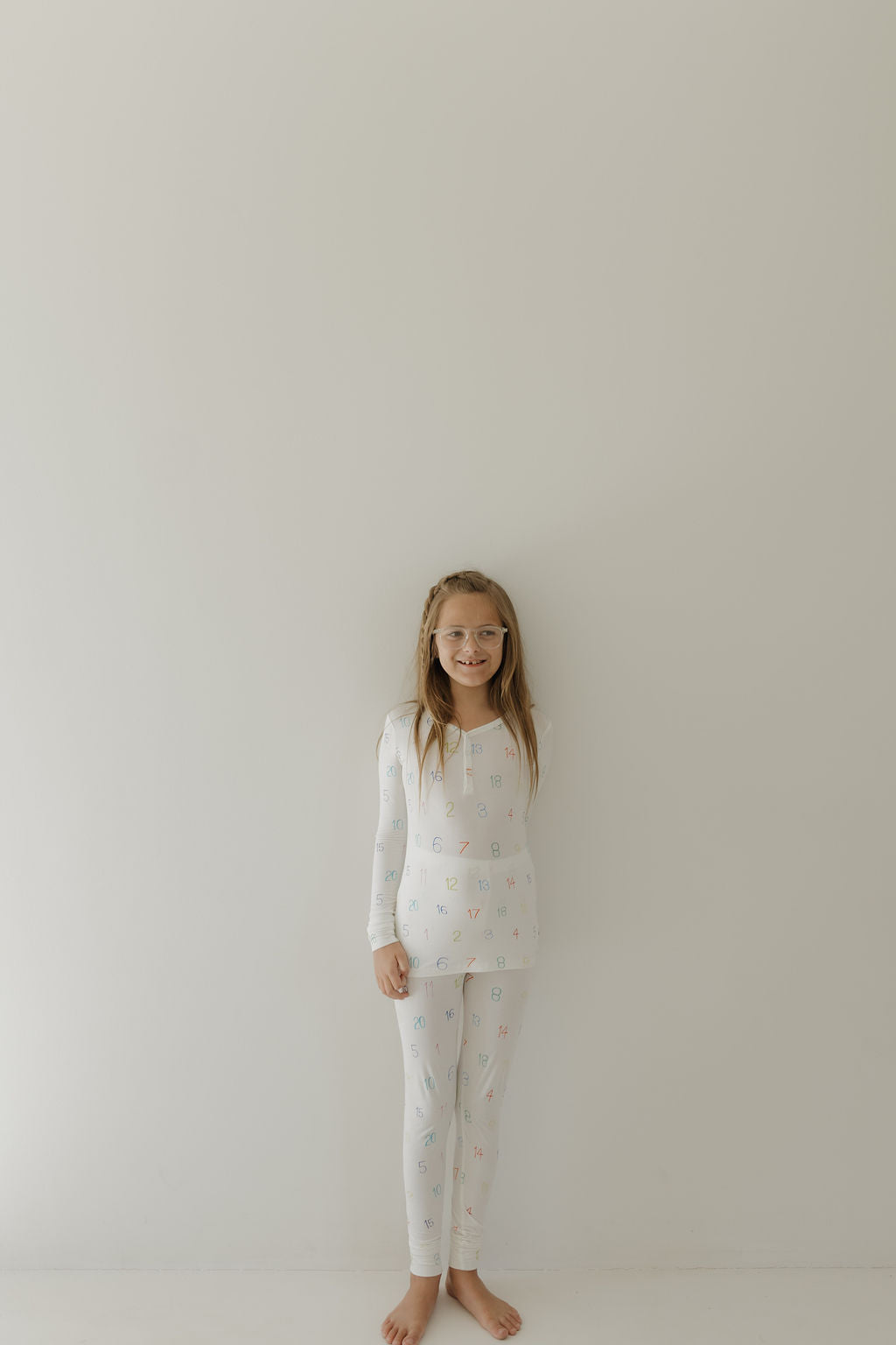 A young girl with long hair and glasses smiles while barefoot against a white wall, wearing forever french baby's hypo-allergenic Bamboo Two Piece Pajamas by Charli Counts, featuring colorful patterns.