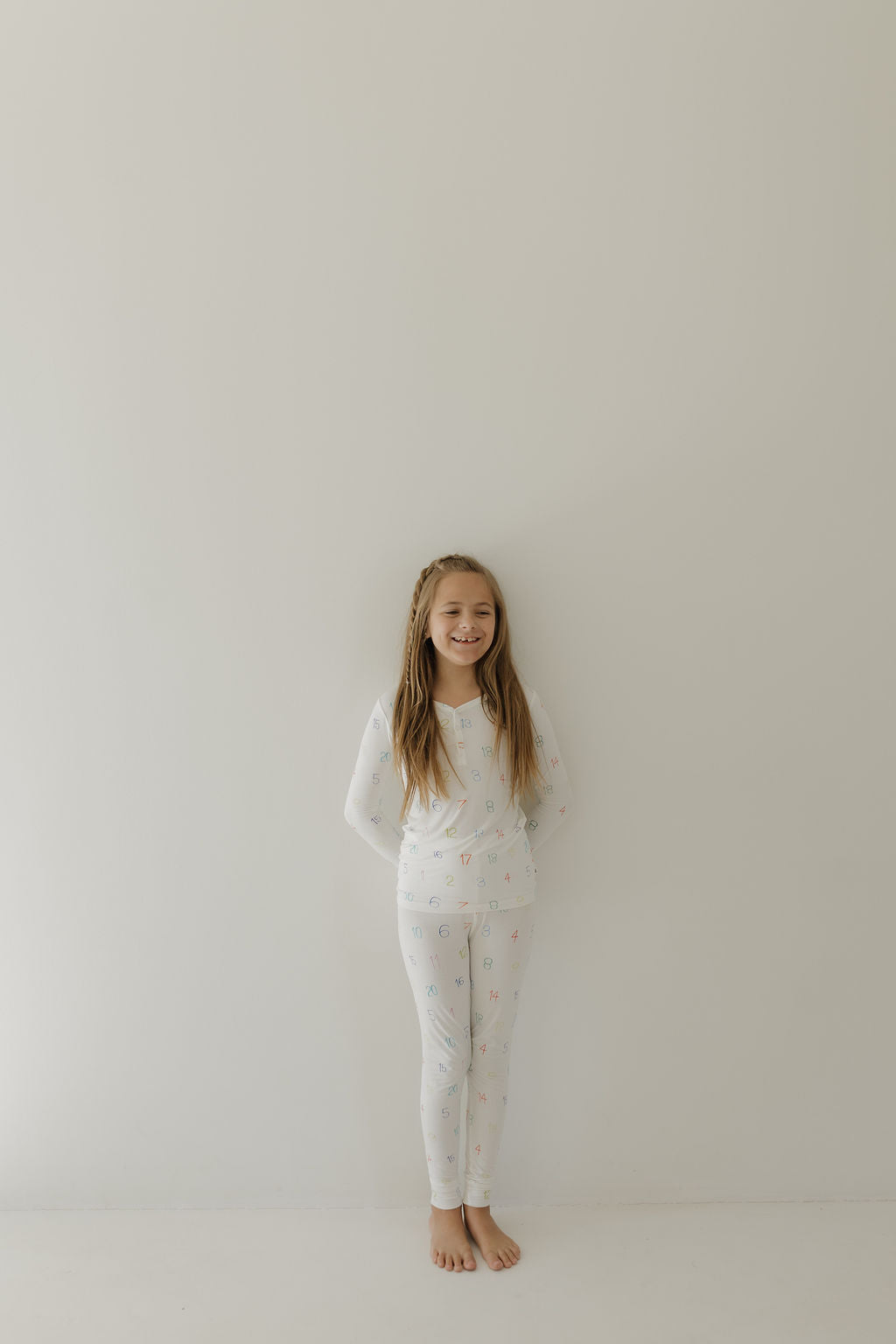 A young girl with long hair stands against a plain white wall, wearing forever french baby Bamboo Two Piece Pajamas in the Charli Counts pattern. She is barefoot and smiling, with her hands playfully tucked behind her back.