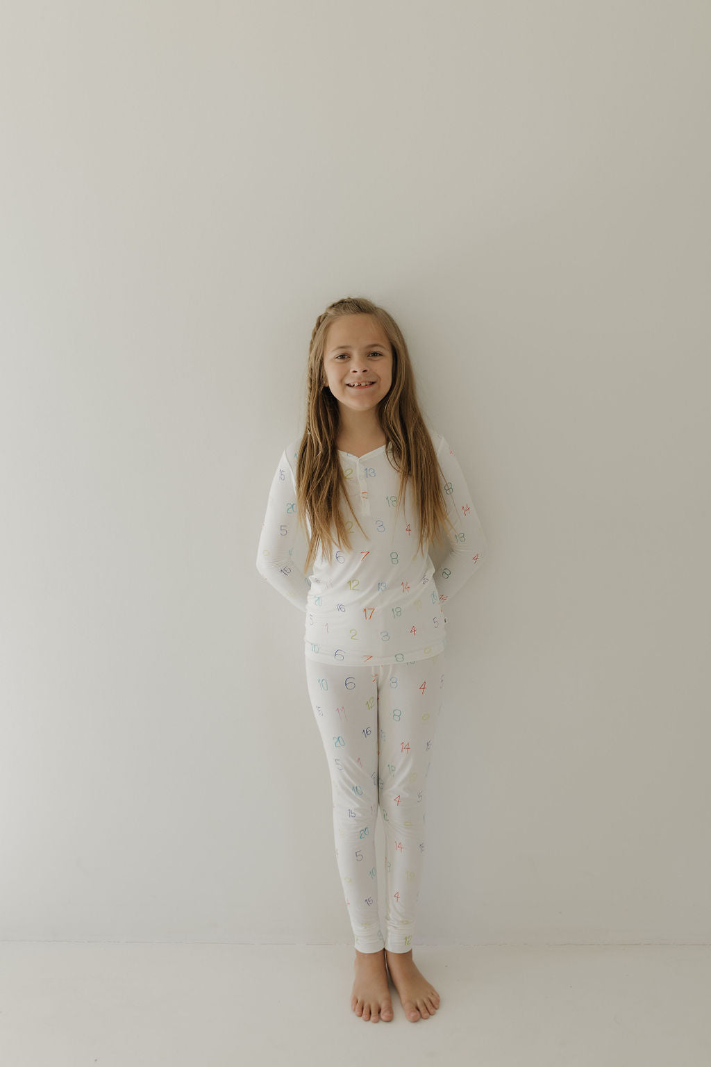 A young girl with long hair stands barefoot against a white wall, smiling warmly as she wears forever french baby Bamboo Two Piece Pajamas | Charli Counts, which are hypo-allergenic and adorned with colorful patterns.