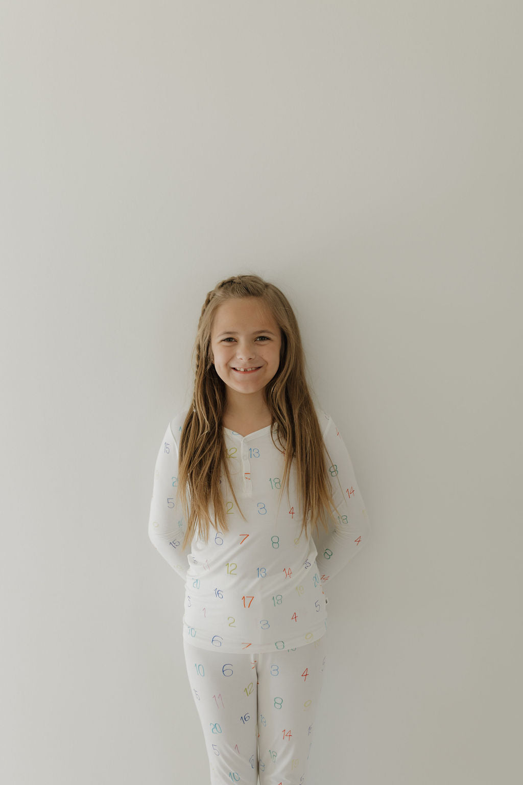 A young girl with long hair smiles against a plain, light-colored wall. Wearing forever french baby's hypo-allergenic Bamboo Two Piece Pajamas | Charli Counts adorned with colorful numbers, she radiates joy and subtly supports autism awareness.