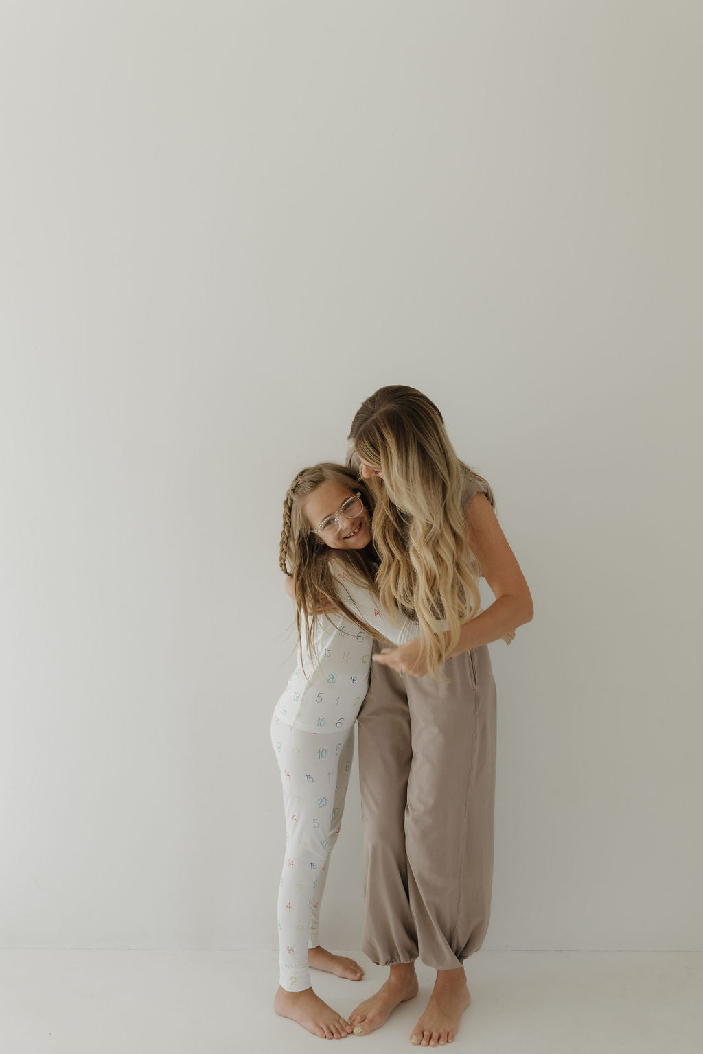 A barefoot woman with long hair embraces a young girl against a white background. The girl wears "Bamboo Two Piece Pajamas" in light colors and patterns from forever french baby, while the woman sports earthy-toned pants.