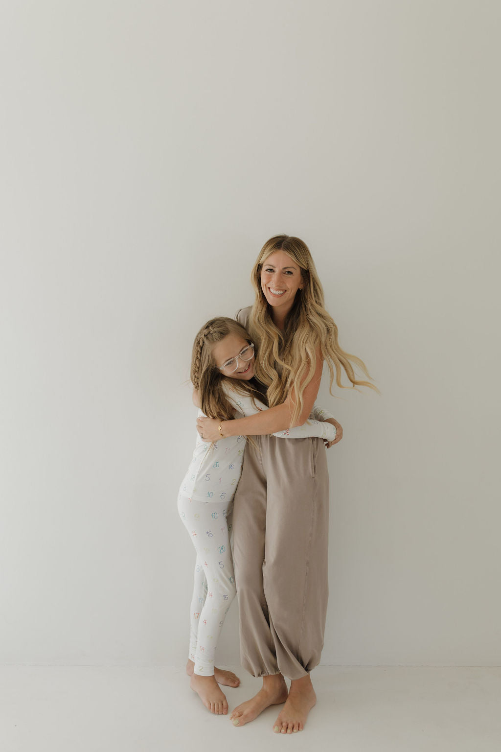 In a bright room, a woman with long hair and a young girl with glasses share a warm hug. The woman wears sleeveless bamboo pajamas, while the smiling girl is cozy in her Charli Counts Bamboo Two Piece Pajamas by forever french baby. Both barefoot, they radiate comfort and warmth.