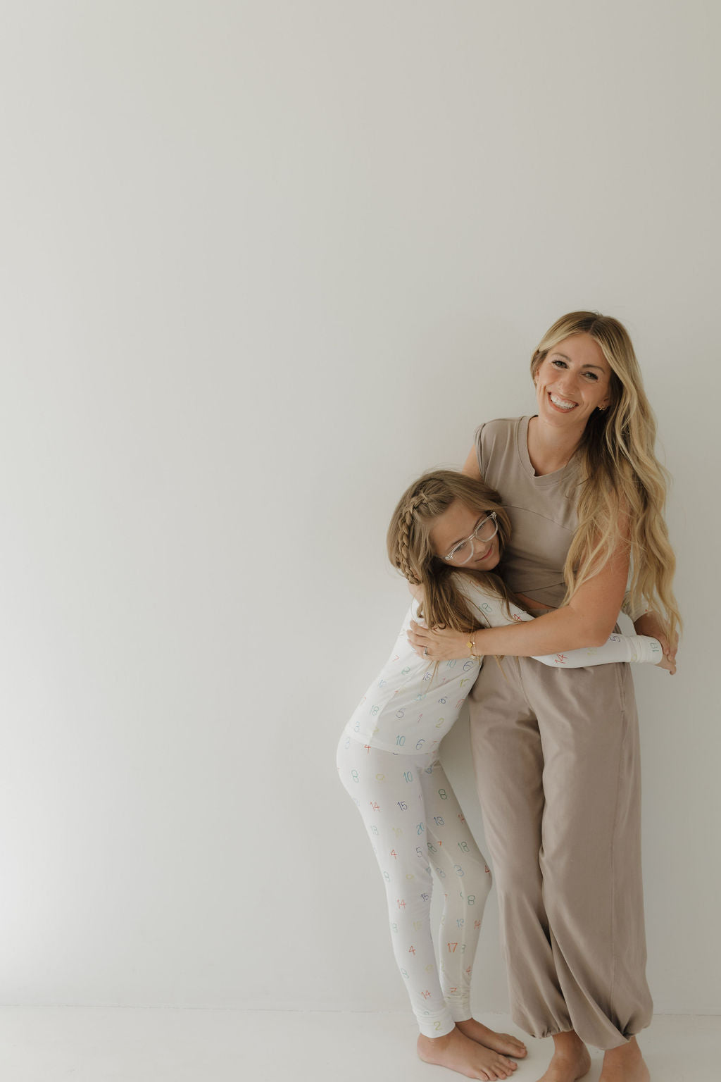 A woman with long, wavy hair smiles as a young girl in glasses and braided hair hugs her. The girl is wearing Bamboo Two Piece Pajamas from the Charli Counts line by forever french baby. The woman sports a taupe outfit, and they both exude warmth against a plain white background.