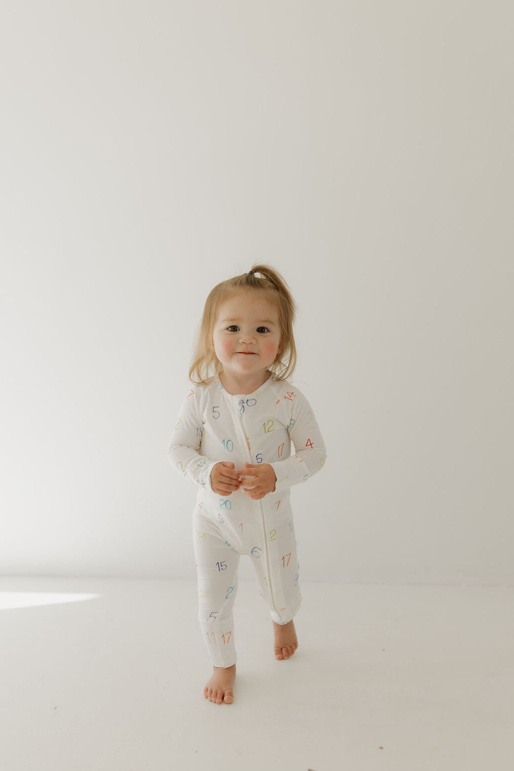 A light brown-haired toddler stands barefoot on a white floor, smiling gently at the camera. They wear forever french baby's Bamboo Zip Pajamas | Charli Counts, made from hypoallergenic bamboo fabric and adorned with multicolored numbers, against a plain white background.