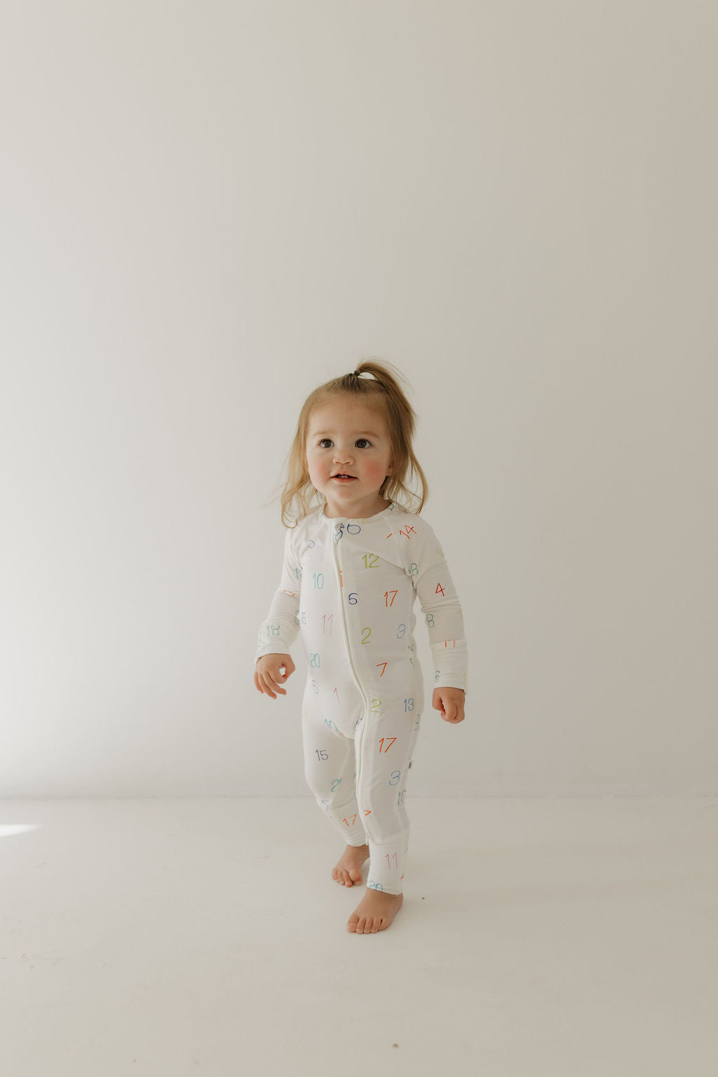 A light brown-haired toddler stands barefoot on a white floor, wearing a forever french baby Bamboo Zip Pajamas | Charli Counts. The outfit is hypo-allergenic bamboo clothing with colorful numbers, and the child looks slightly to the side against a plain white background.