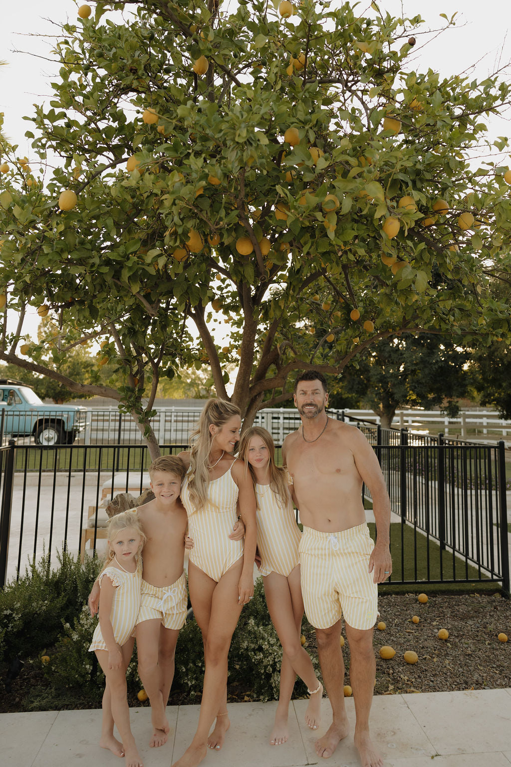 A family of five in matching yellow and white striped lolo webb 2 swimsuits, featuring Men Board Short | Sun Rays, poses by a lemon tree near a black fence, with a blue truck and greenery in the background.