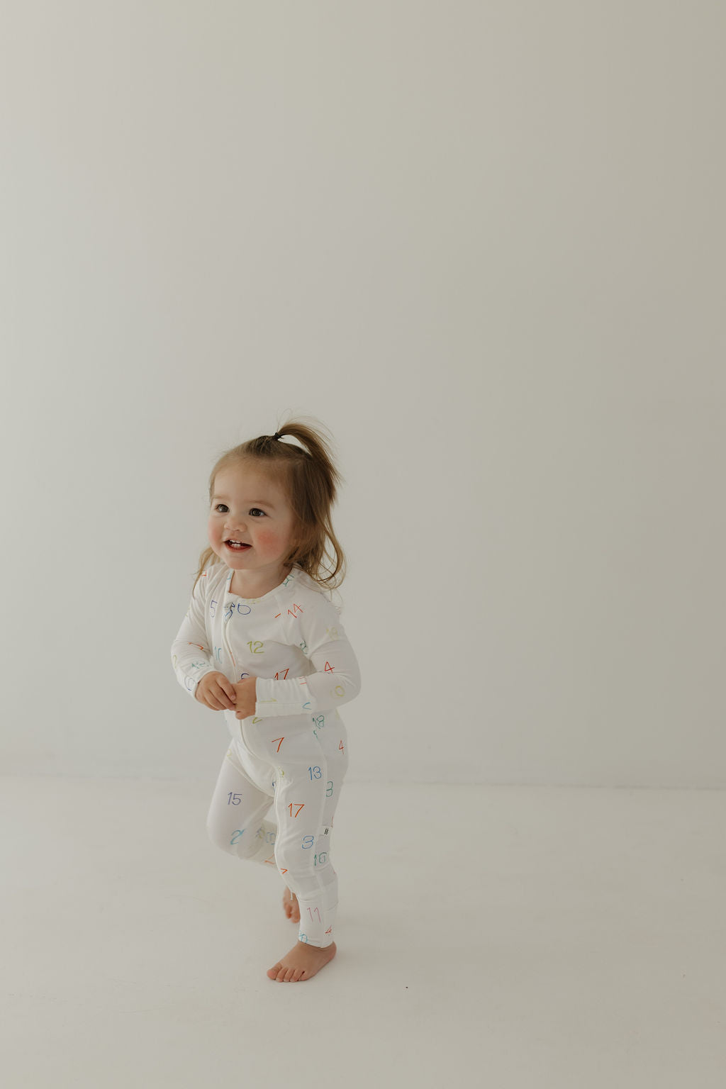 A joyful young child with a ponytail stands barefoot against a light background, wearing forever french baby's Bamboo Zip Pajamas | Charli Counts, a hypo-allergenic white onesie adorned with colorful numbers.