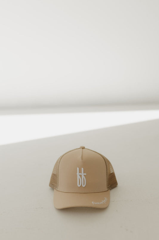 A Children's Trucker Hat in tan with a white logo from forever french baby sits on a light surface. The background subtly shifts from light to shadow, enhancing the minimalistic vibe that embodies the Forever French Baby aesthetic.