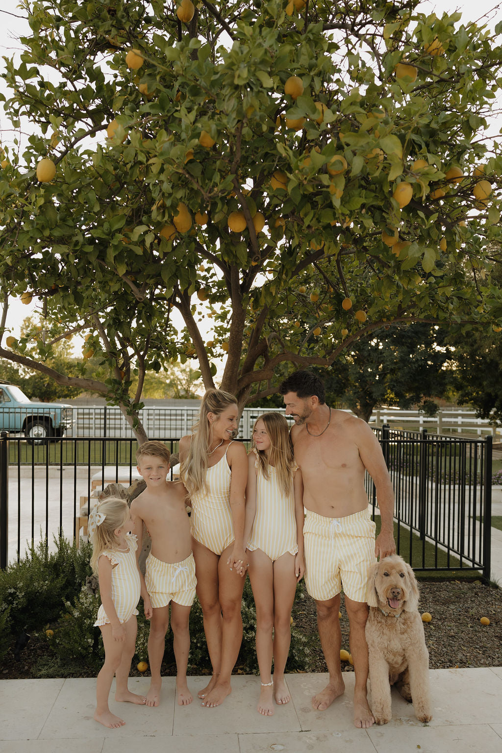 A family of five and their dog smile barefoot beneath a lemon tree, all wearing matching lolo webb 2 Child Board Short | Sun Rays. A black fence and blue vehicle are visible in the background.
