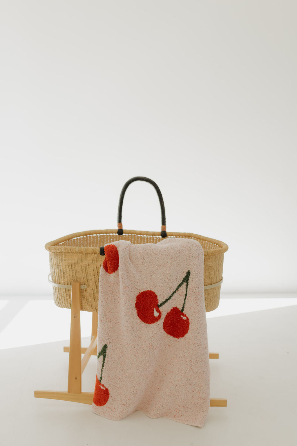 A woven baby bassinet on a wooden stand against a light background features the "Plush Blanket | Cherry Orchard" by forever french baby, an OEKO Standard 100 light pink blanket with red cherries draped over the side for a pop of color and extra softness.