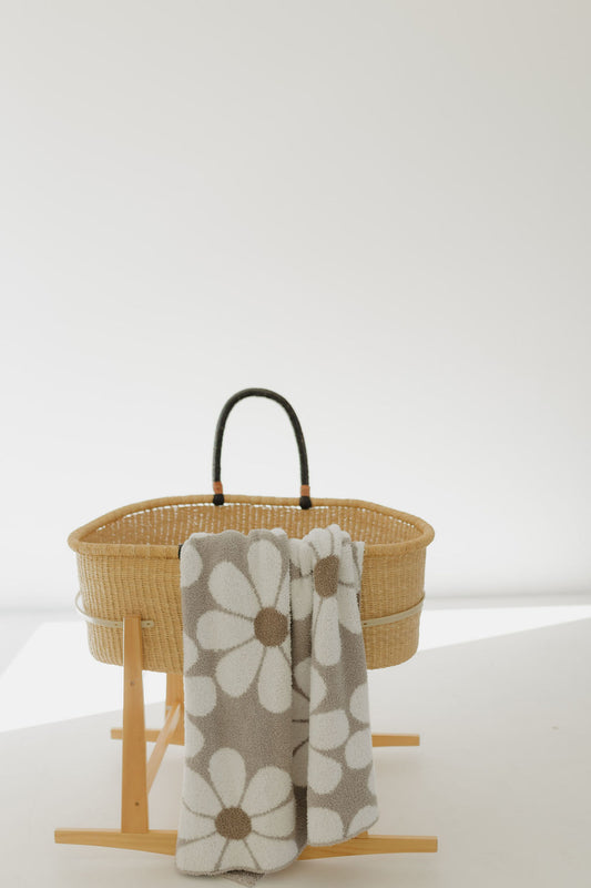 A wicker bassinet with a dark handle is placed on a wooden stand against a white backdrop. Draped over the side is forever french baby's Plush Blanket | Darlin', Oeko-tex certified for safety, featuring large white and brown daisy patterns. Bright light creates soft shadows around the setup.