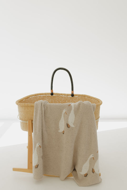 A woven bassinet with black handles sits on a wooden stand. Draped over the edge is the forever french baby Plush Blanket | Silly Goose, crafted from Oeko-Tex certified microfiber and adorned with charming white goose prints.