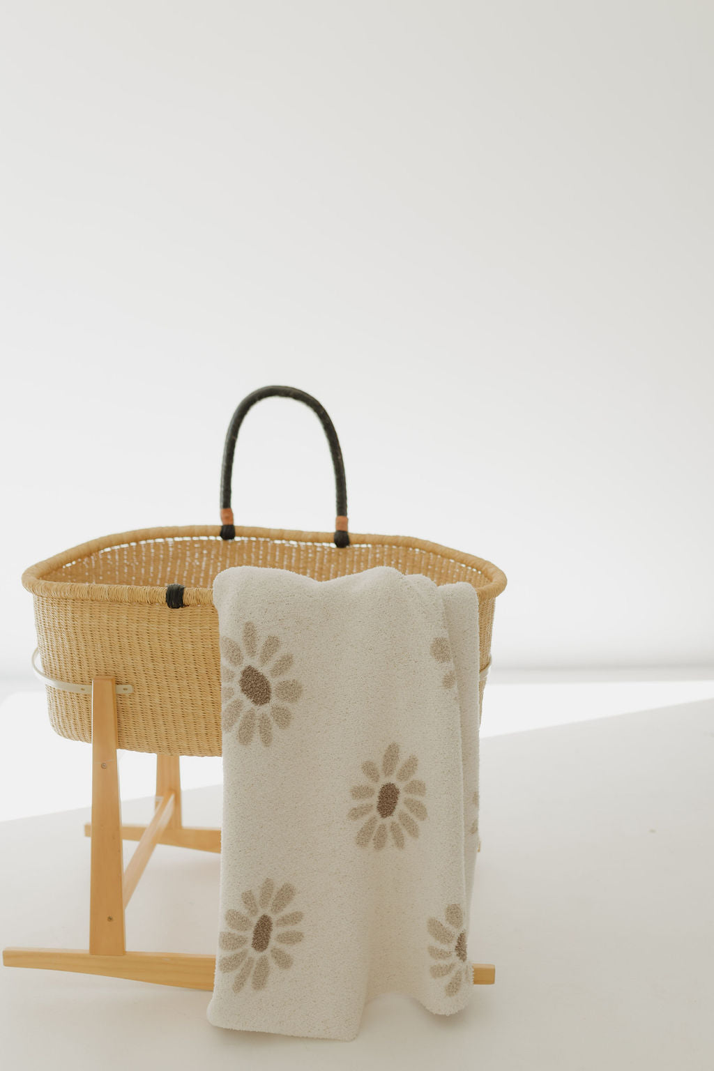 A woven baby bassinet with dark handles rests on a wooden stand. Draped over the side is the "Plush Blanket | Daisy Fields" by Forever French Baby, crafted from OEKO-TEX certified microfiber feather yarn with a daisy pattern. The setting is a minimalist, bright white space.