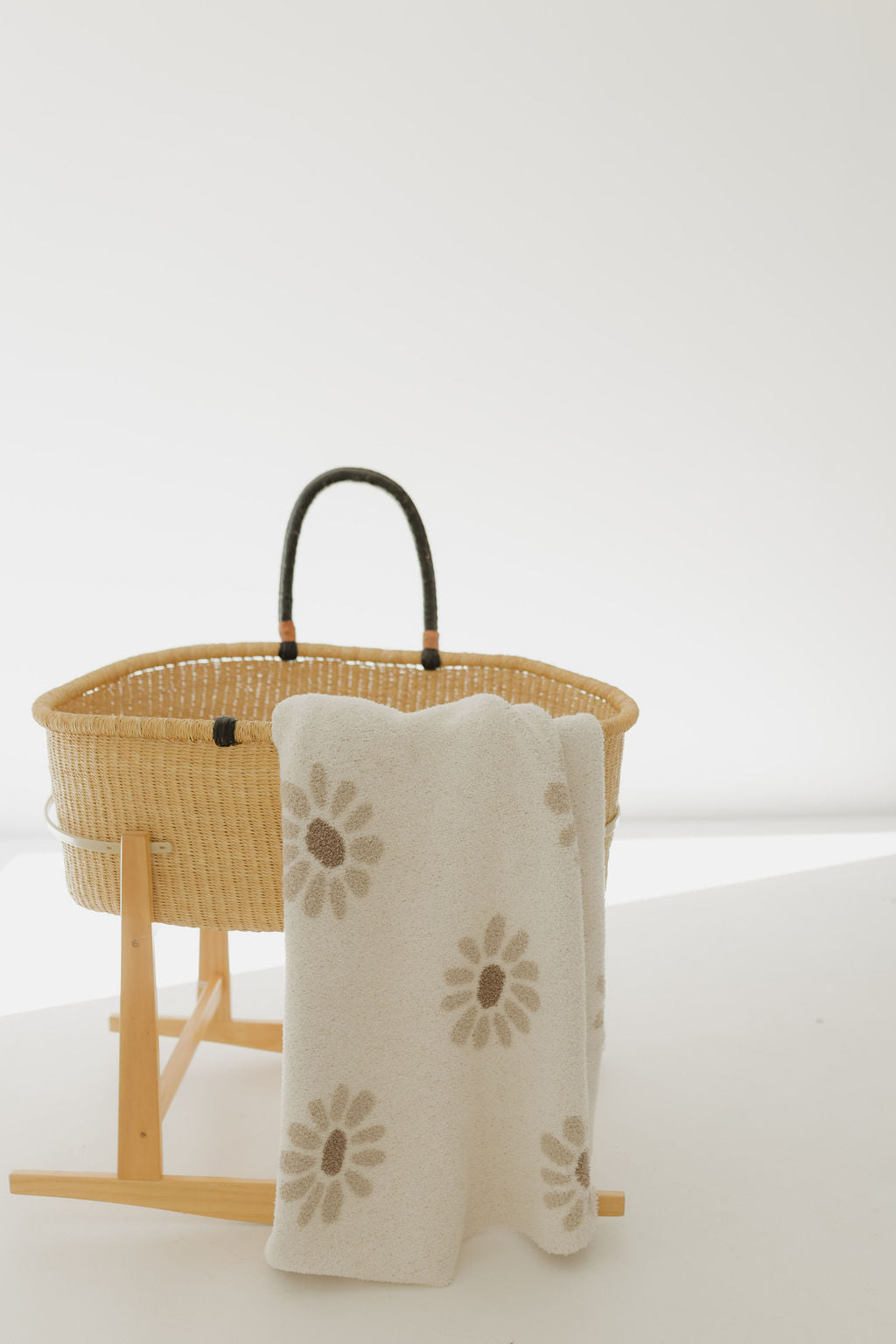 A wicker baby bassinet with black handles on a wooden stand is draped with the "Plush Blanket | Daisy Fields" by forever french baby. This white blanket, featuring brown daisy patterns and made from OEKO-TEX certified materials, stands out against the minimal and brightly lit background.