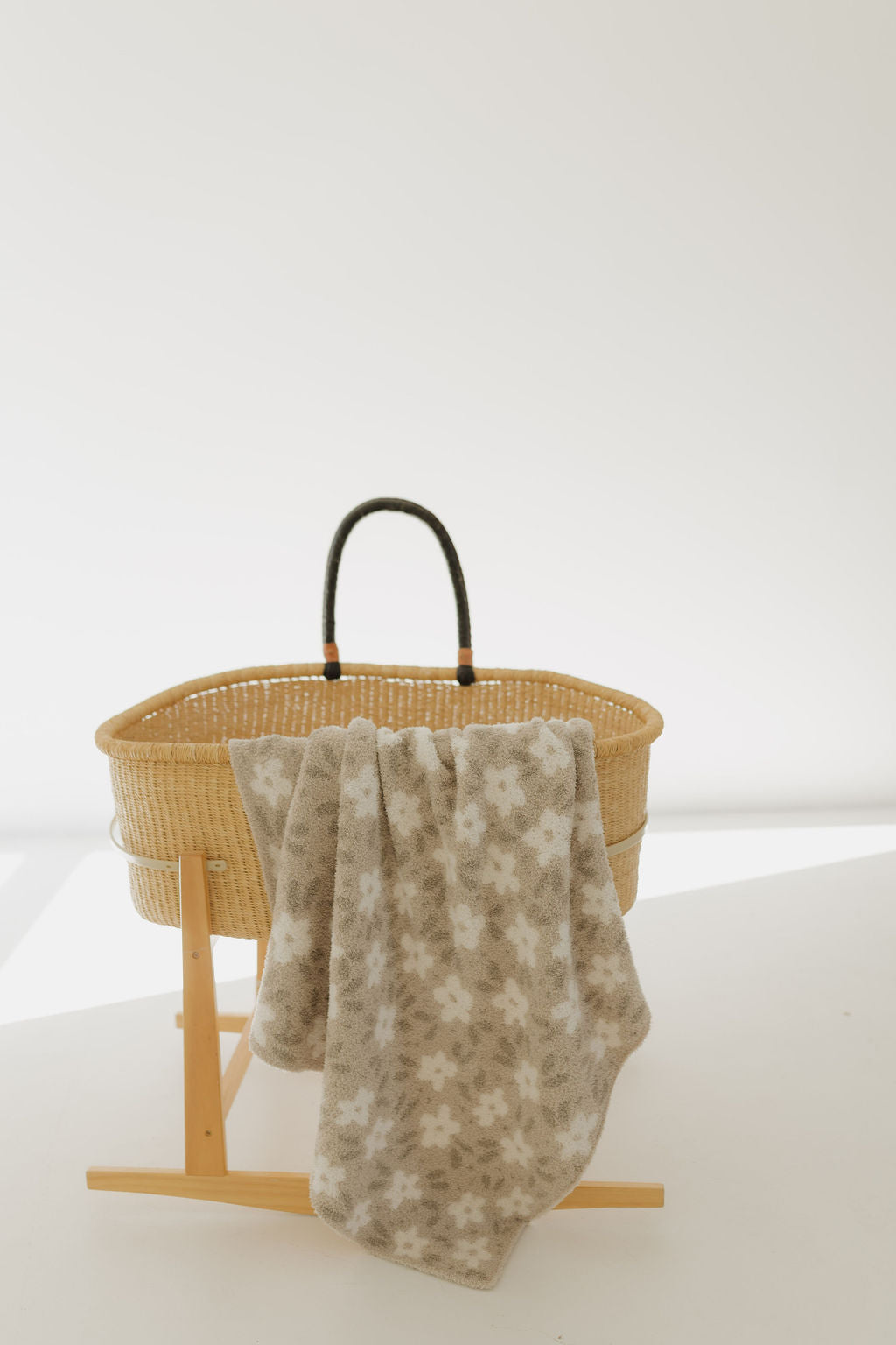 A wicker bassinet with a black handle stands on a wooden frame in a bright room. Draped over it is a plush, Oeko-tex certified blanket, the "French Gray Floral" from forever french baby. The background is plain and softly lit.