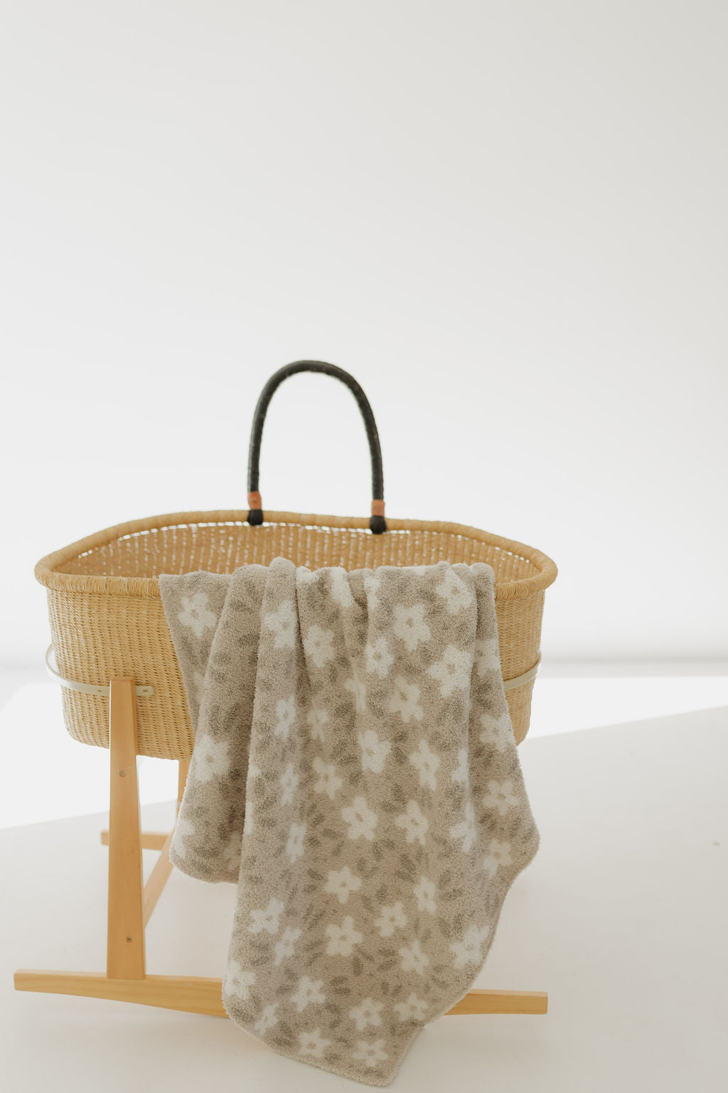A baby bassinet with a wicker basket and wooden stand is shown, adorned with the "Plush Blanket | French Gray Floral" by forever french baby. The background is softly lit, enhancing the serene and cozy ambiance.