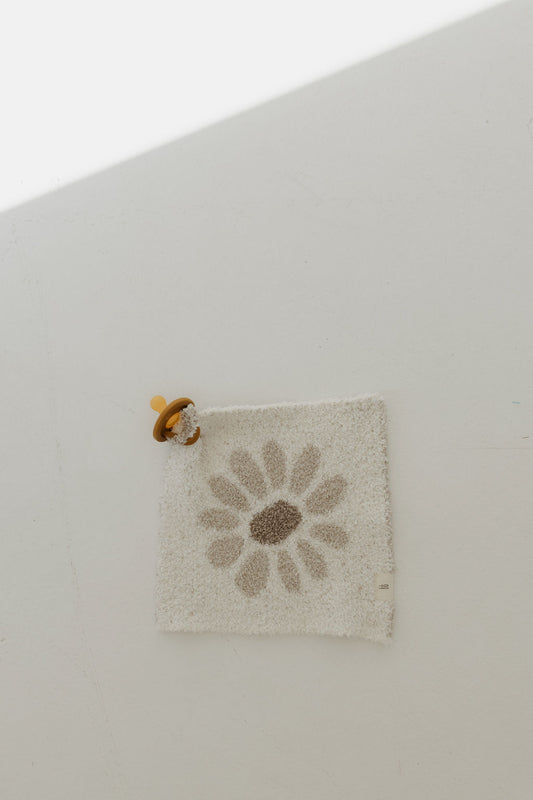 The Lovey | Daisy Fields by forever french baby lies on a light surface. A brown pacifier with a yellow ring is attached to one corner of the square white mat. Crafted from microfiber feather yarn, it glows as sunlight streams in at an angle.