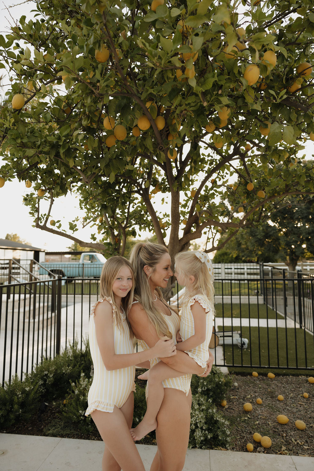 A woman and two young girls wear lolo webb 2 Women's One Piece Swimsuit | Sun Rays in yellow and white stripes, smiling together under a lemon tree in a sunny backyard scattered with lemons.