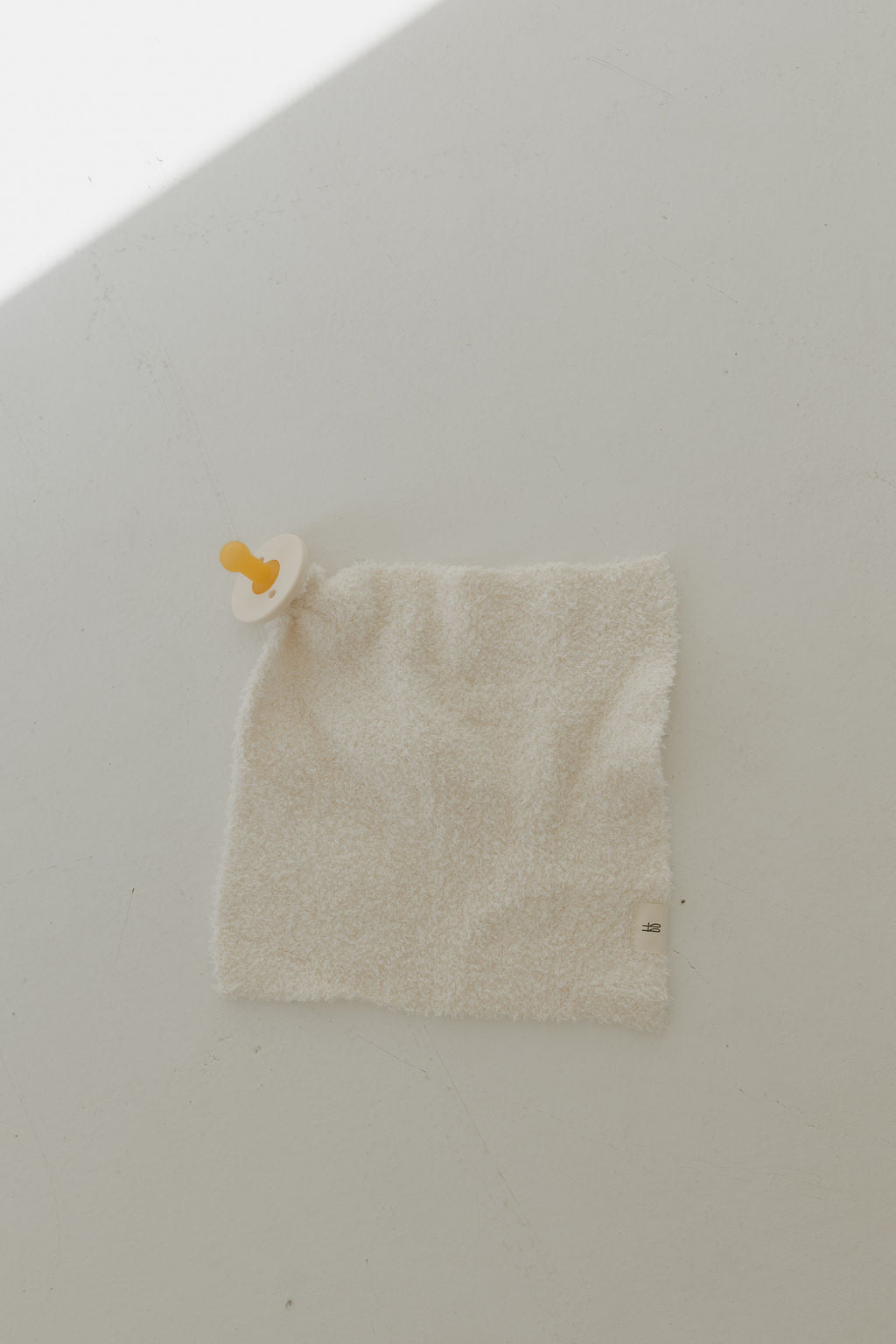 A cream Lovey by Forever French Baby, made of textured microfiber feather yarn with an attached pacifier, rests on a smooth, light surface. A sliver of light crosses the upper left corner, casting subtle shadows.