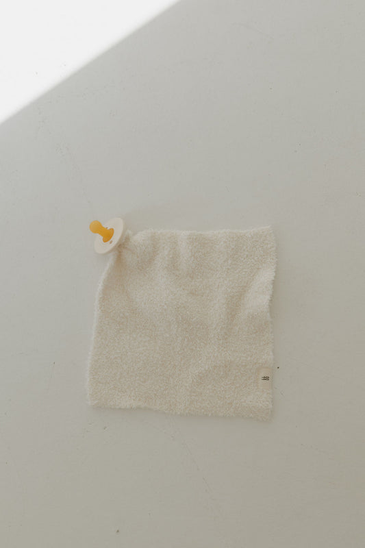 A cream Lovey by Forever French Baby, made of textured microfiber feather yarn with an attached pacifier, rests on a smooth, light surface. A sliver of light crosses the upper left corner, casting subtle shadows.