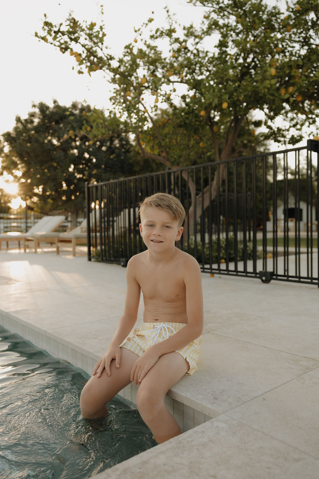 A young boy in the lolo webb 2 Child Board Short | Sun Rays sits poolside with his feet in the water, smiling at the camera. Behind him, a black fence and trees set the scene for this classic outdoor moment.