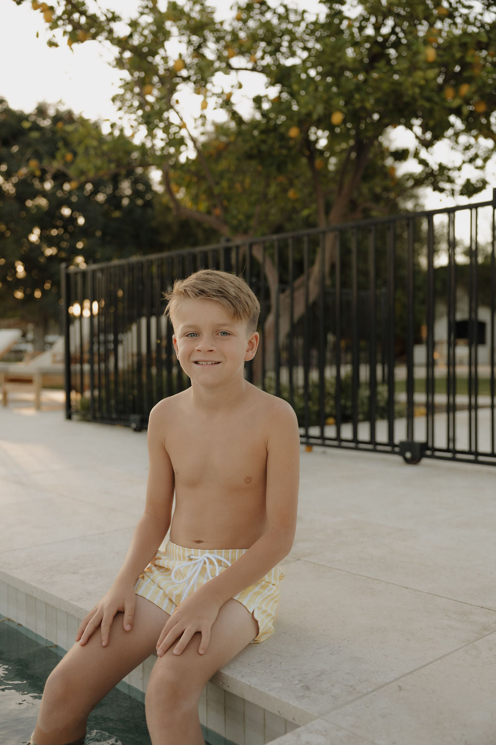 A young boy wearing lolo webb 2’s Child Board Short | Sun Rays smiles poolside. A black metal fence and trees frame the relaxed outdoor setting in the background.