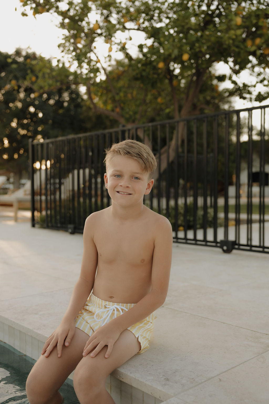 A young boy with short blond hair smiles at the camera while sitting by a pool, wearing lolo webb 2's Child Board Short | Sun Rays. Behind a black fence, trees with green leaves are visible.