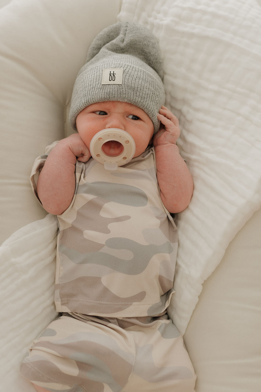 A baby in a hypo-allergenic Bamboo Short Set by forever french baby, featuring a camo pattern, lies on a soft white blanket. The child wears a gray beanie, holds it with one hand while having a pacifier in its mouth and gazing upwards.