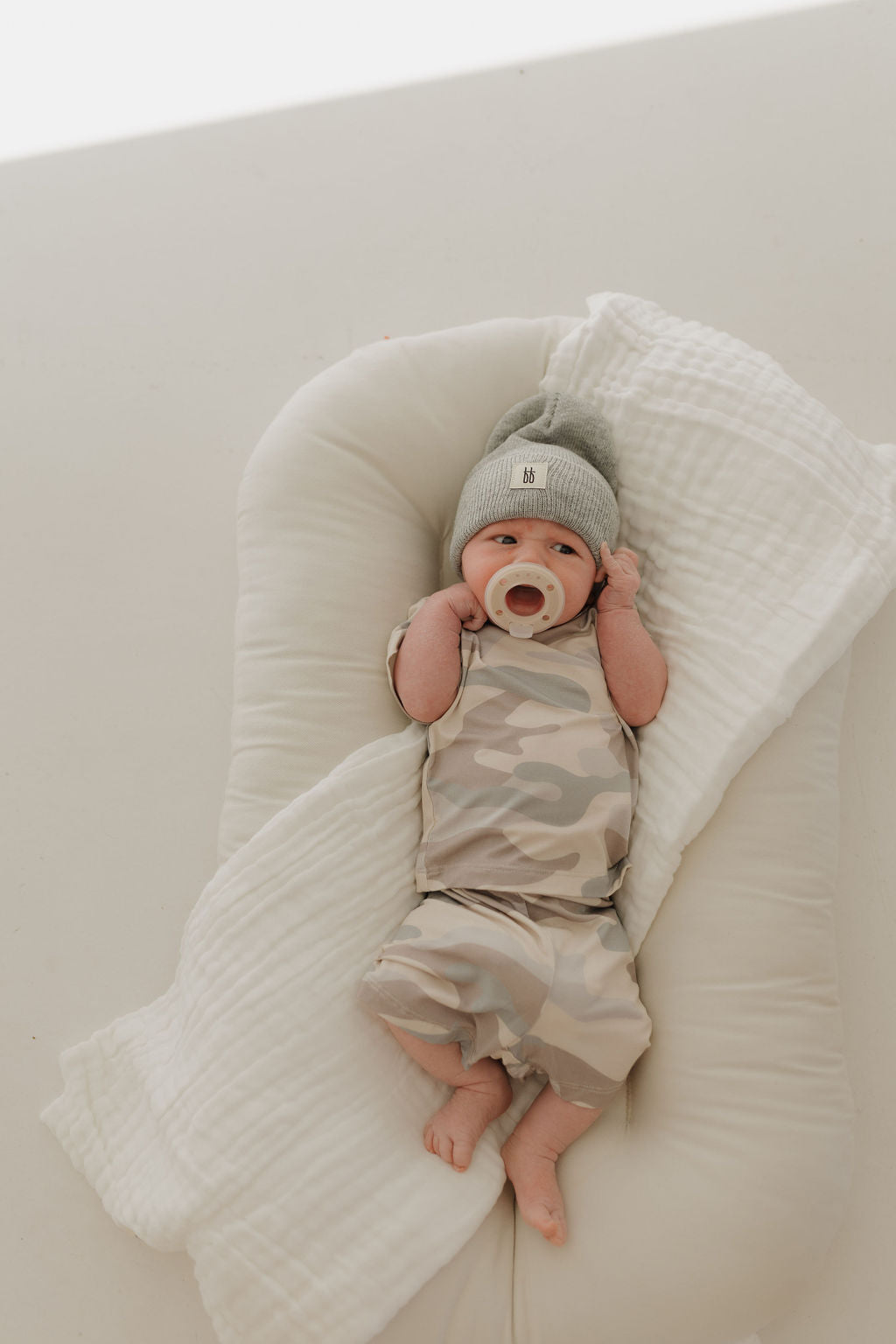 A baby in a forever french baby Bamboo Short Set | Camo and a gray-green beanie lies on a soft, white cushion, with a pacifier in its mouth, wrapped in breathable, hypo-allergenic muslin cloth for maximum comfort.