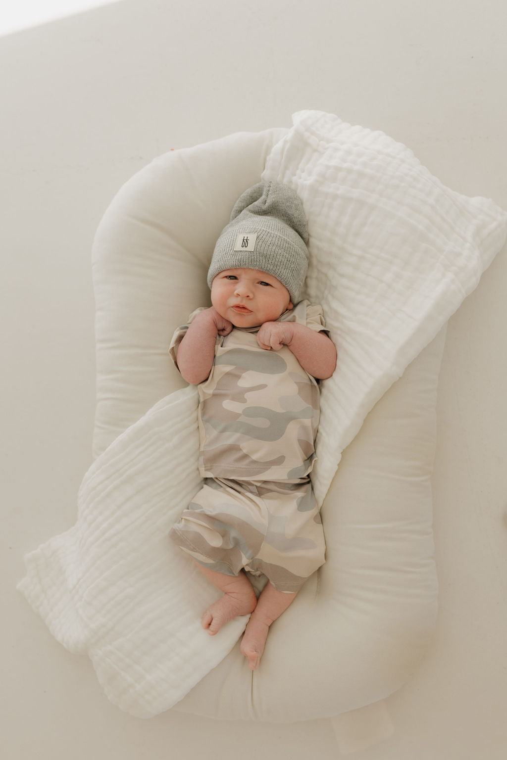 A baby in a Bamboo Short Set | Camo by forever french baby peacefully rests on a white cushioned mat, wearing a gray beanie and wrapped in a light-colored hypoallergenic blanket, with tiny fists near their face.