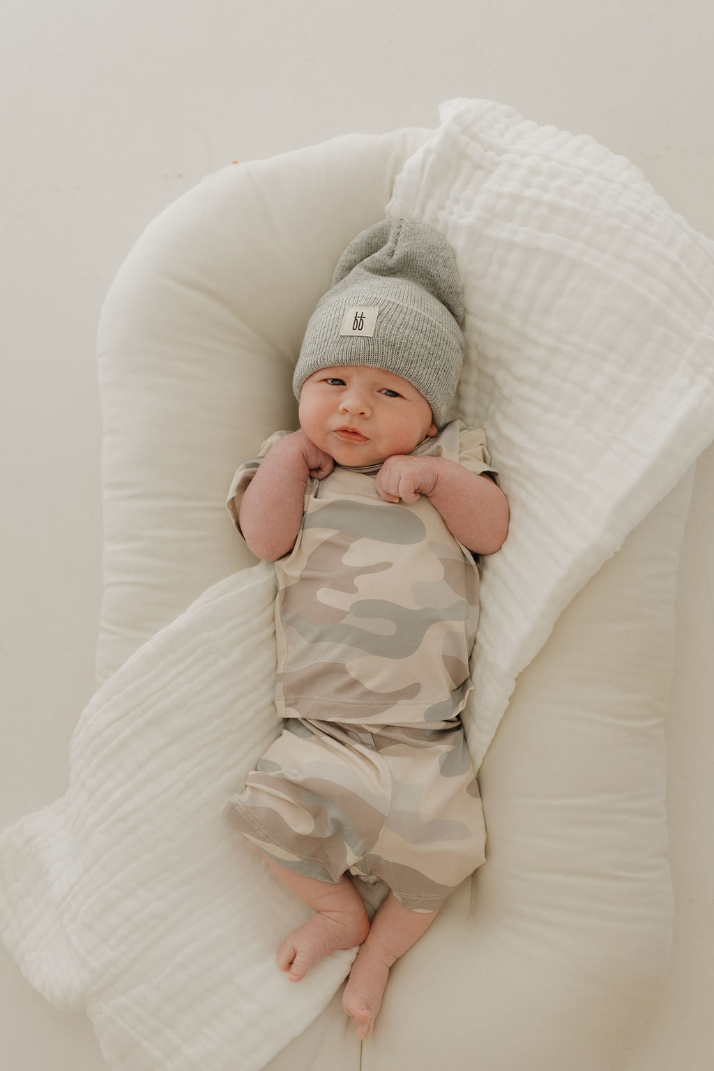 A baby wearing the Bamboo Short Set in Camo from forever french baby, rests on a white, cushioned blanket. The gray beanie complements the breathable sleepwear perfectly. The baby looks up with hands near its face, lying in a relaxed pose.