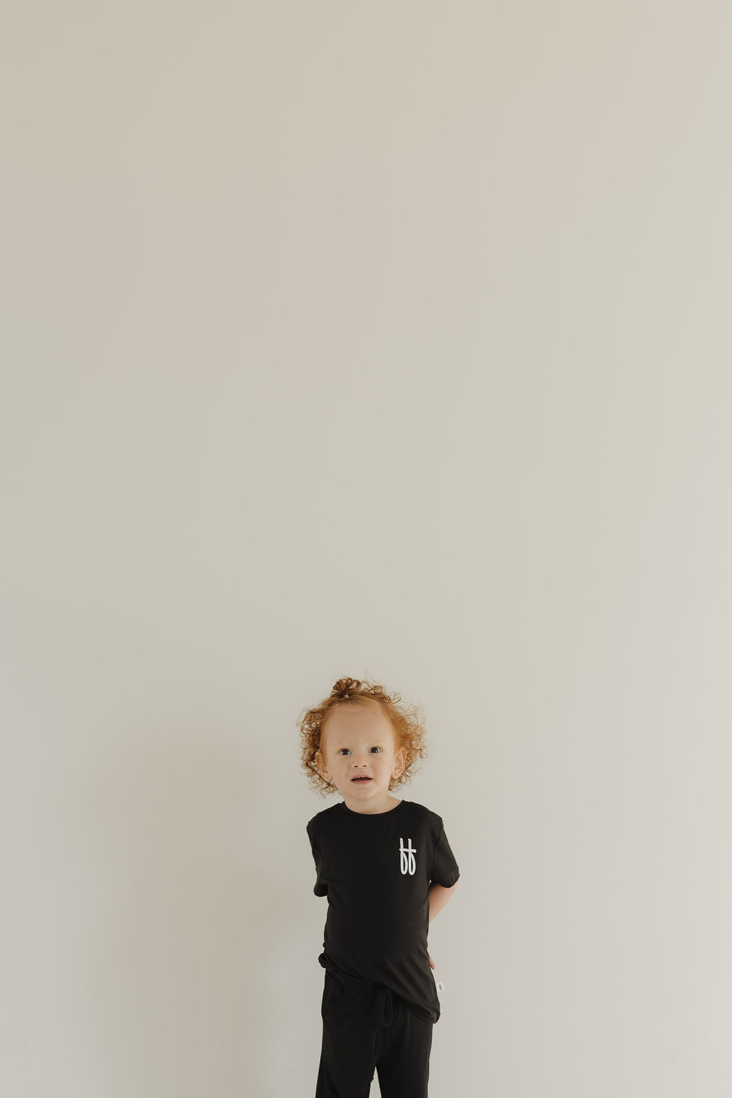 A curly-haired toddler stands against a plain, light wall, wearing the forever french baby Black ff Signature Bamboo Lounge Set made from hypo-allergenic bamboo. They gaze directly at the camera.