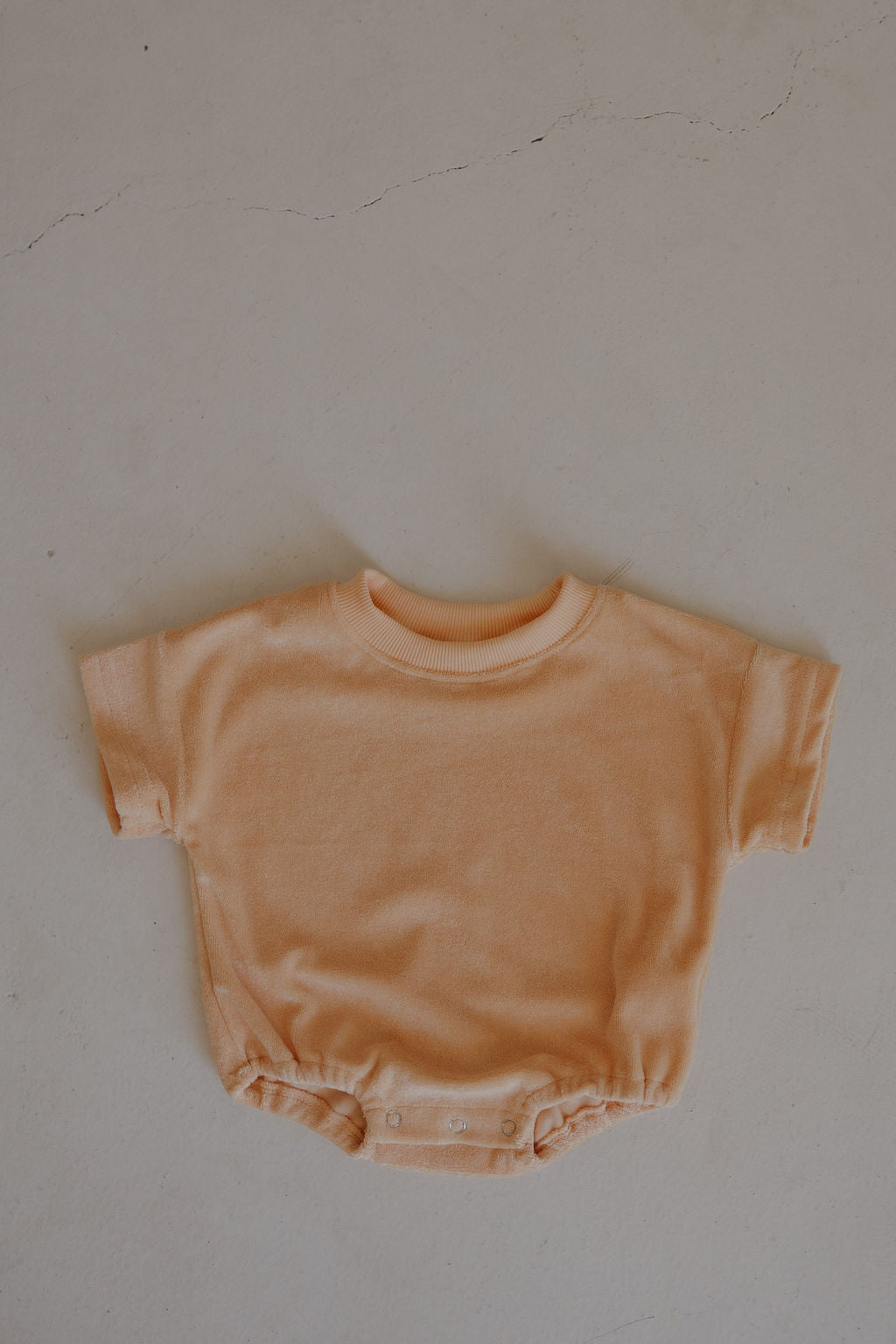 Sherbert peach Terry T-Shirt Romper by forever french baby, featuring short sleeves and snap buttons at the bottom for easy diaper changes, displayed flat on a light, textured surface.