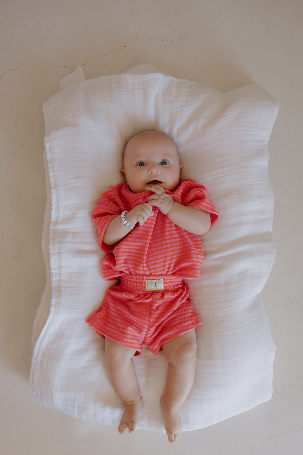 A baby lies on a white blanket, wearing the forever french baby Child Terry Short Set in Strawberry, with a small bracelet and hands near their mouth.