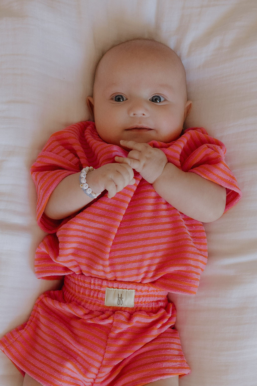 A baby in the forever french baby Child Terry Short Set | Strawberry lies on a white blanket, gazing upward with wide eyes and clasped hands—a sweet family moment, highlighted by a tiny beaded bracelet.