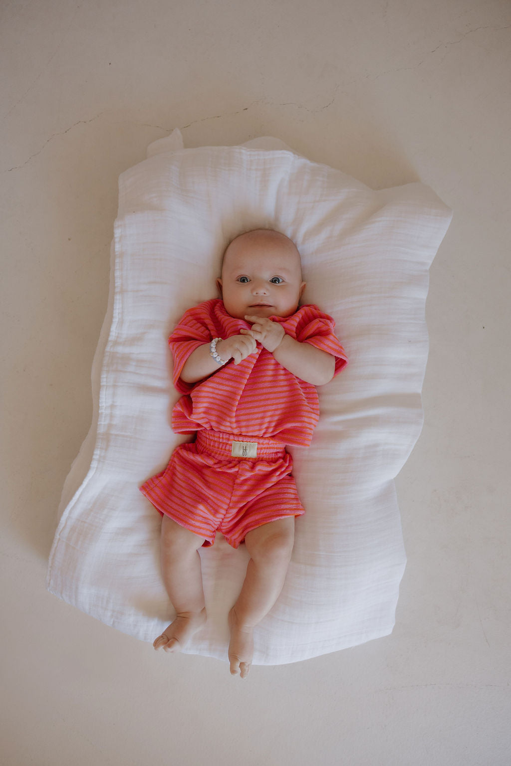 A baby wears the forever french baby Child Terry Short Set in Strawberry, lying on a white blanket with hands near their mouth against a light beige floor—perfect for capturing precious family moments.