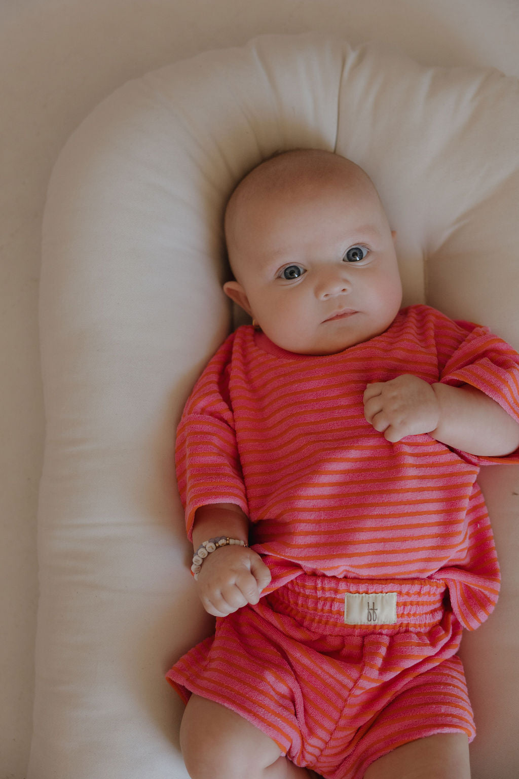 A baby lies on a soft, white cushion, wearing the forever french baby Child Terry Short Set | Strawberry and a beaded bracelet, gazing wide-eyed at the camera.