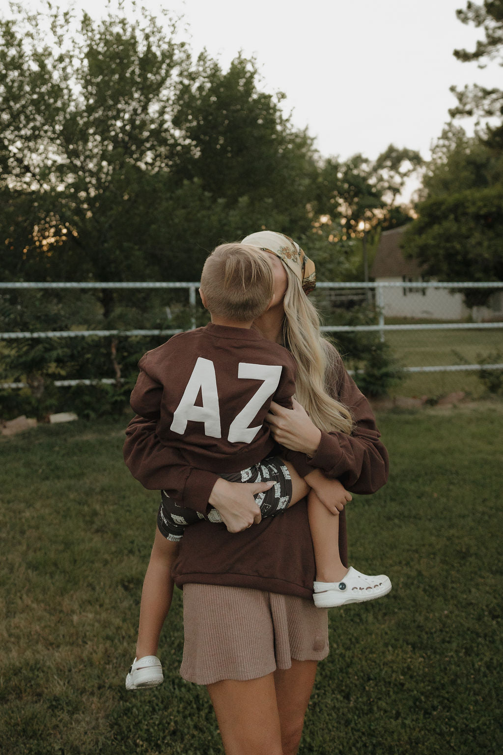 A woman in a headband and kids cotton sweatshirt stands in a grassy yard, holding a young boy wearing the forever french baby Child Sweatshirt | ff Arizona. Behind them, trees and a fence set the scene for cozy matching moments.